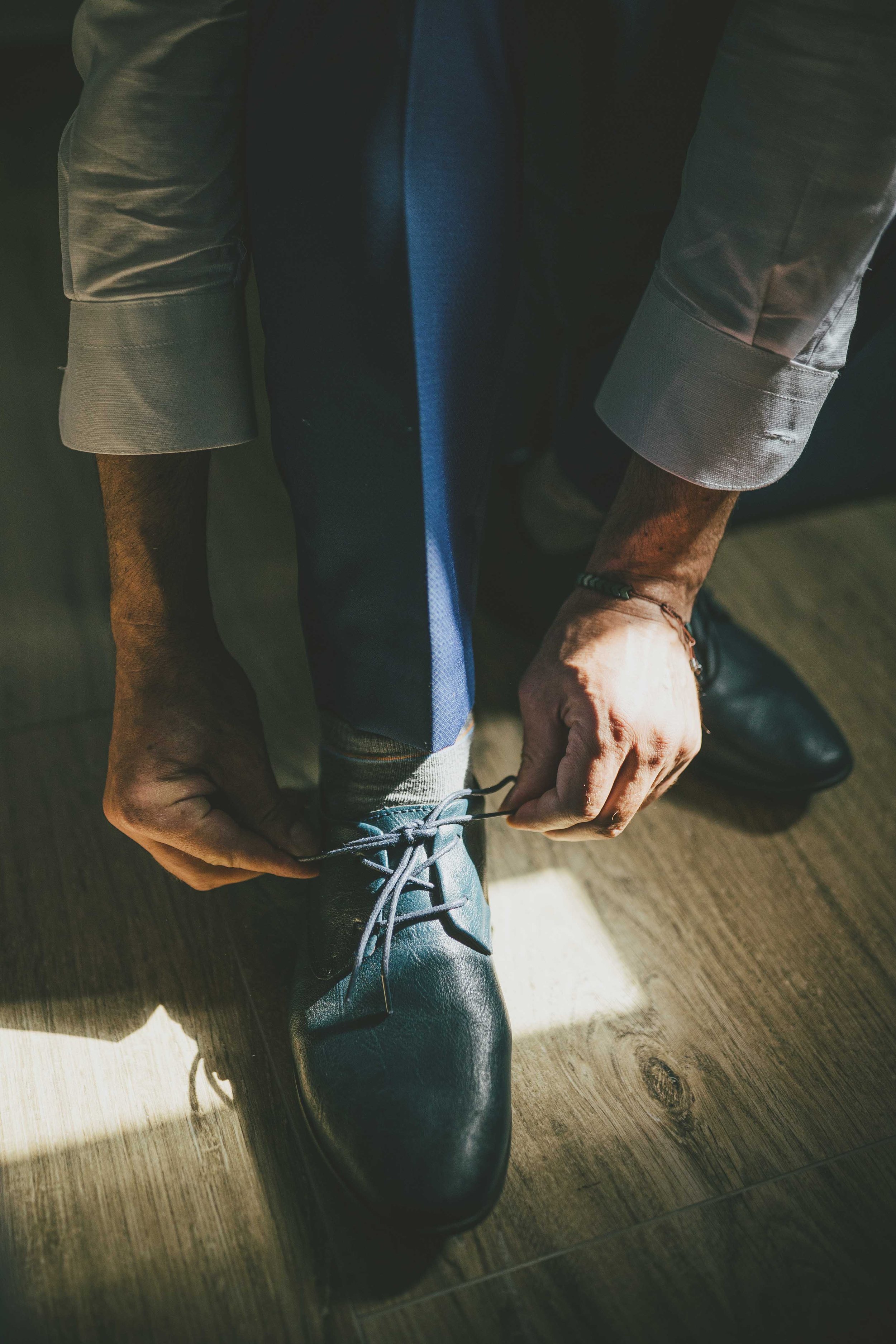 Eine Person bindet ihre schwarzen Schuhe, trägt eine graue Jacke und eine blaue Hose, die auf einem Holzboden liegt.