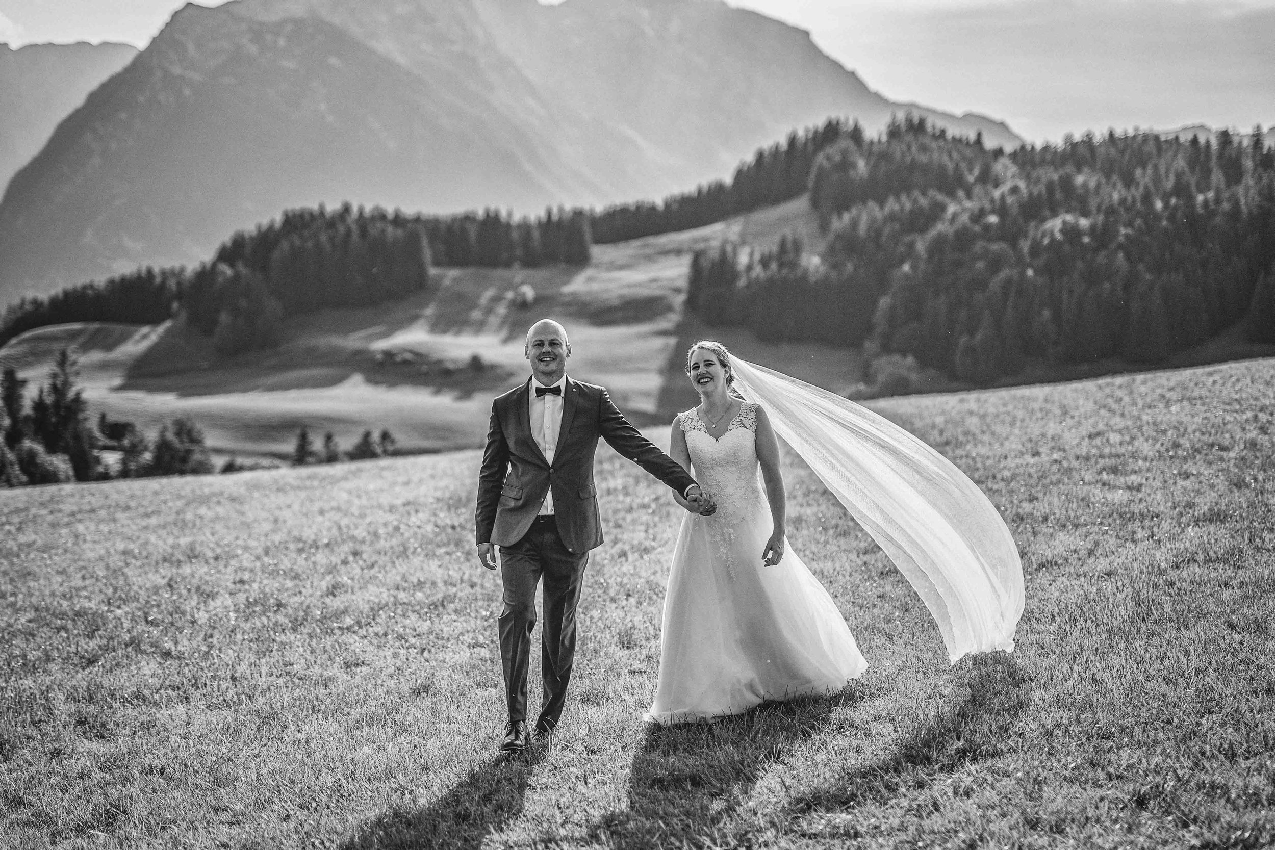 Ein glückliches Brautpaar läuft Hand in Hand über eine Wiese vor Bergen, in Schwarz-Weiß aufgenommen.