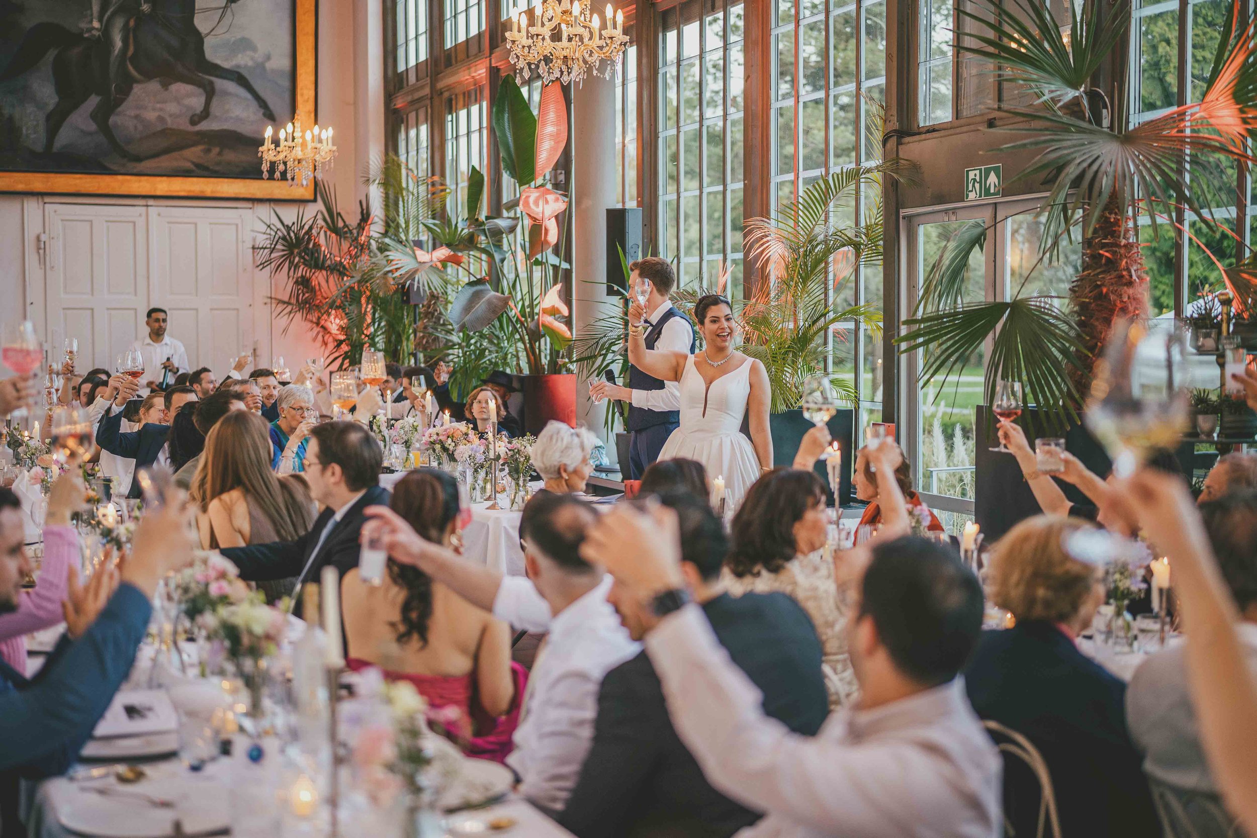 Hochzeitspaar steht in einem festlich dekorierten Raum mit vielen Gästen, die anstoßen, im Hintergrund große Fenster und exotische Pflanzen