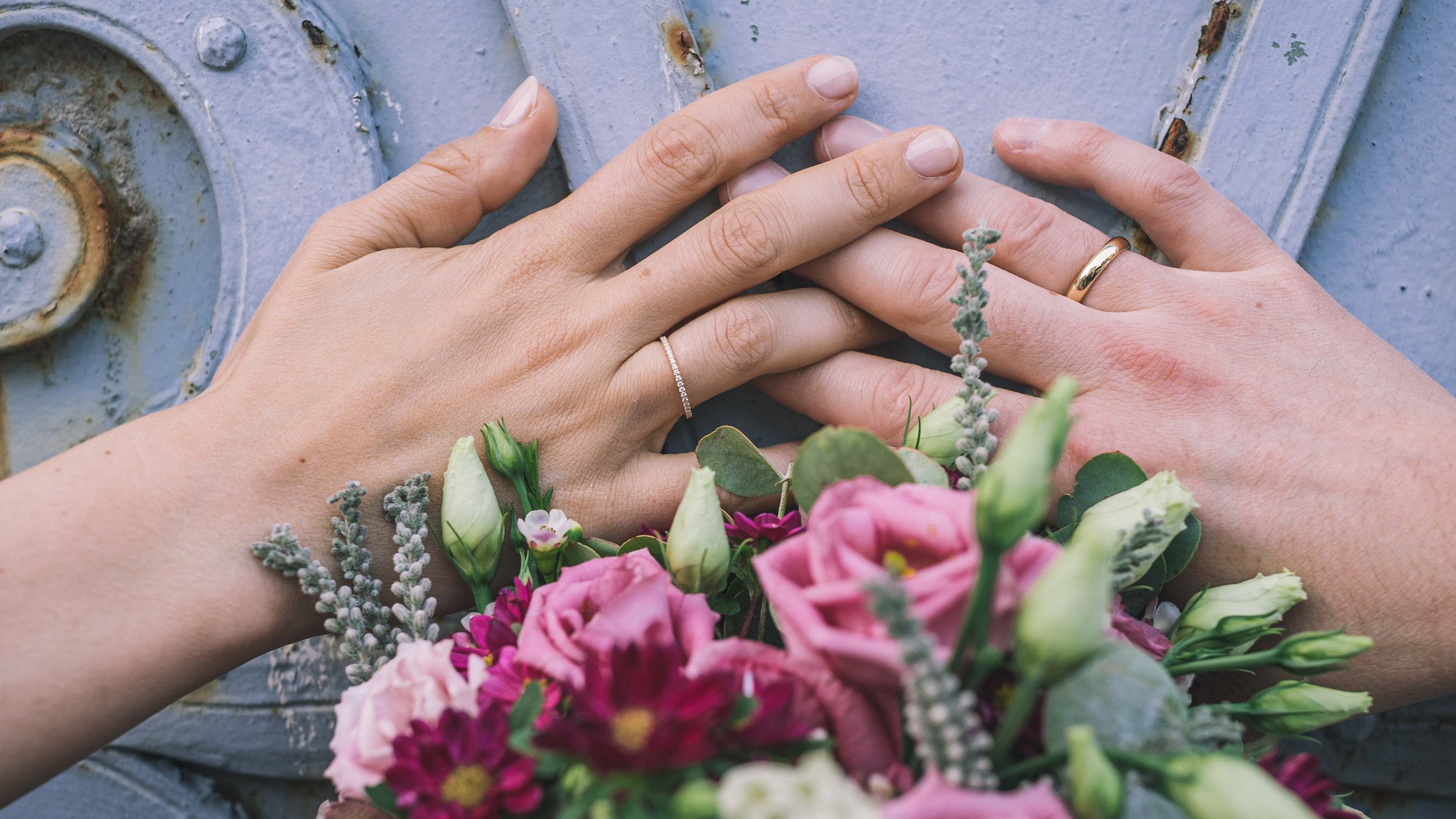 Zwei Hände mit Eheringen liegen auf einer blauen Metallfläche, umgeben von einem Blumenstrauß mit pinken, weißen und lila Blumen.