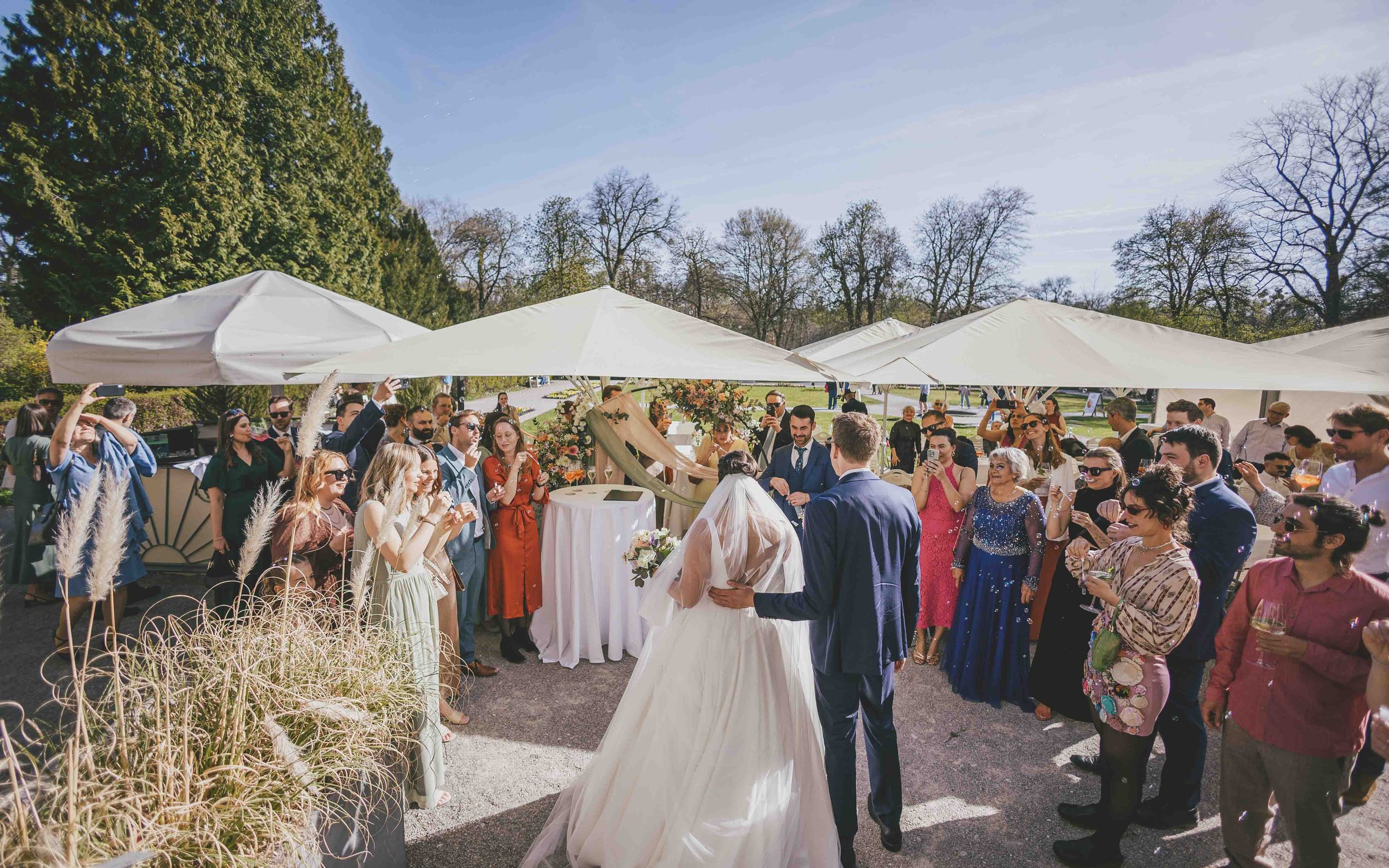 Hochzeit im Freien mit einer Braut in Weiß, einem Bräutigam in Anzug, umgeben von Gästen, die Fotos machen und feiern, unter weißen Sonnenmarkisen, im Grünen bei schönem Wetter.