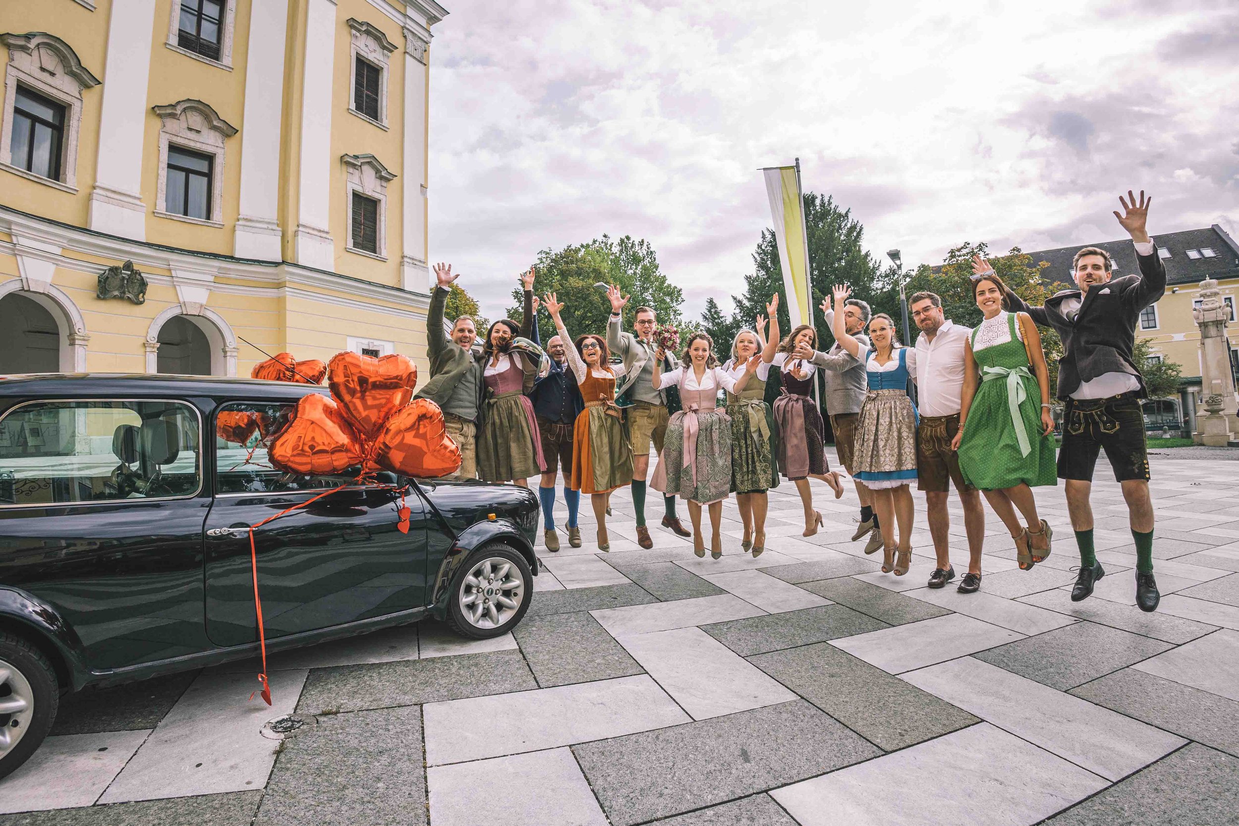 Gruppe von jungen Menschen in traditionellen Trachten beim Jubeln, neben einem schwarzen Auto mit Herzballons, vor einem gelben historischen Gebäude, wahrscheinlich bei einer Hochzeit in Bayern.