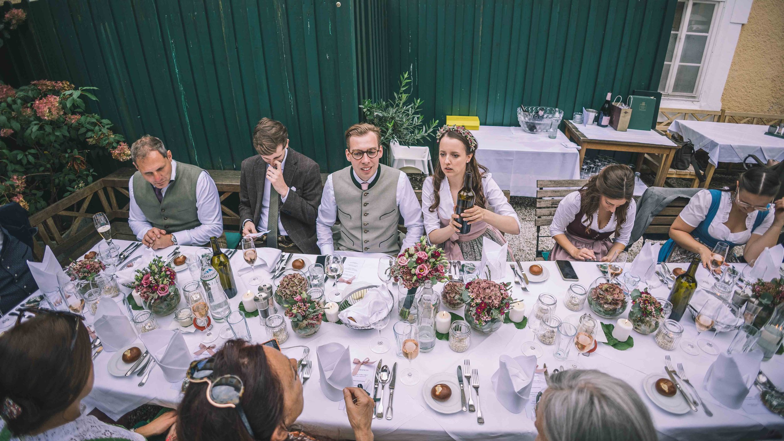 Gruppen von Gästen beim festlichen Essen im Garten, gedeckter Tisch mit Blumen, Kerzen und Gläsern, Personen in traditioneller Kleidung.