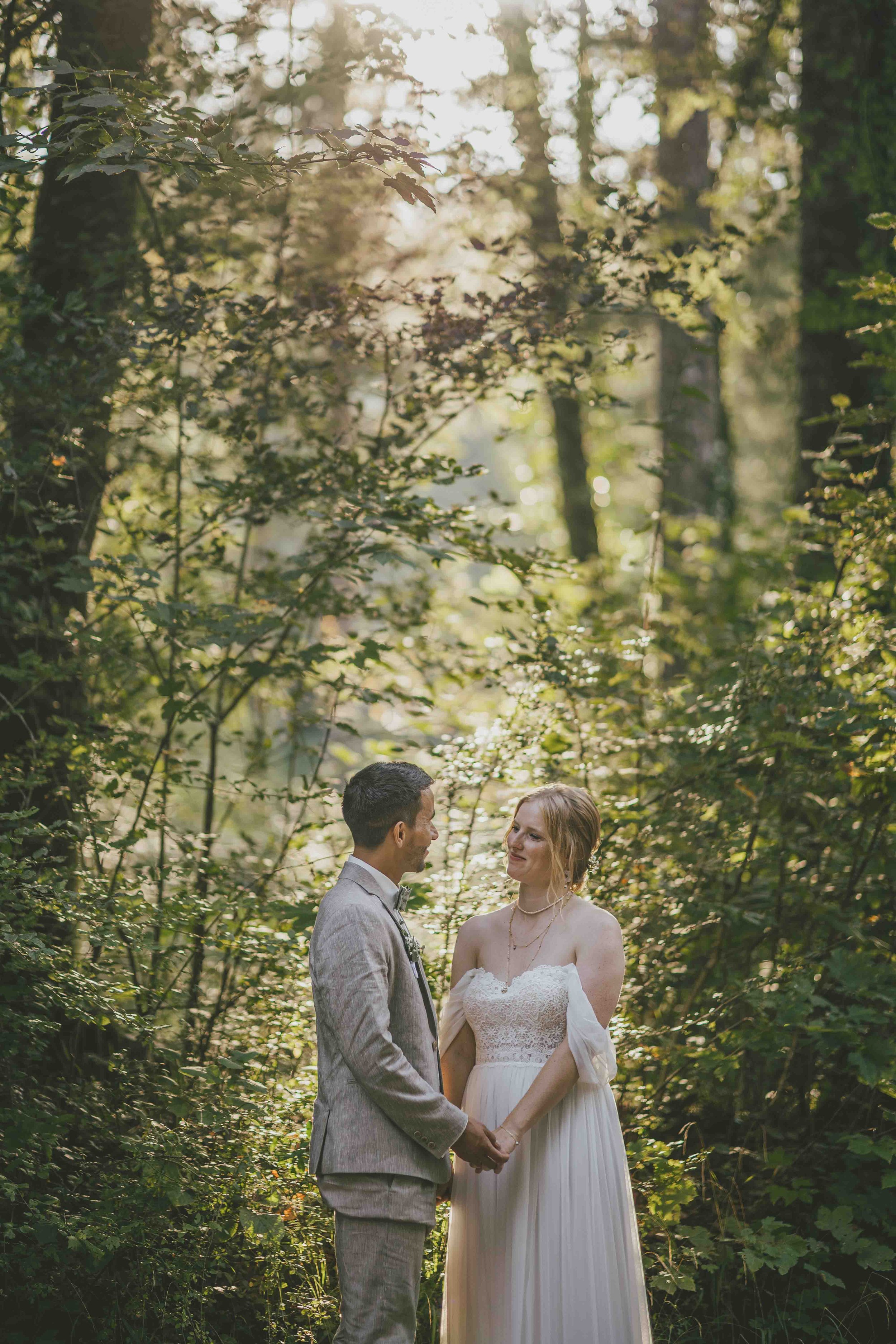 Ein Brautpaar in einem Wald, hält Händchen und schaut sich verliebt an, umgeben von grünen Bäumen und Sonnenlicht.