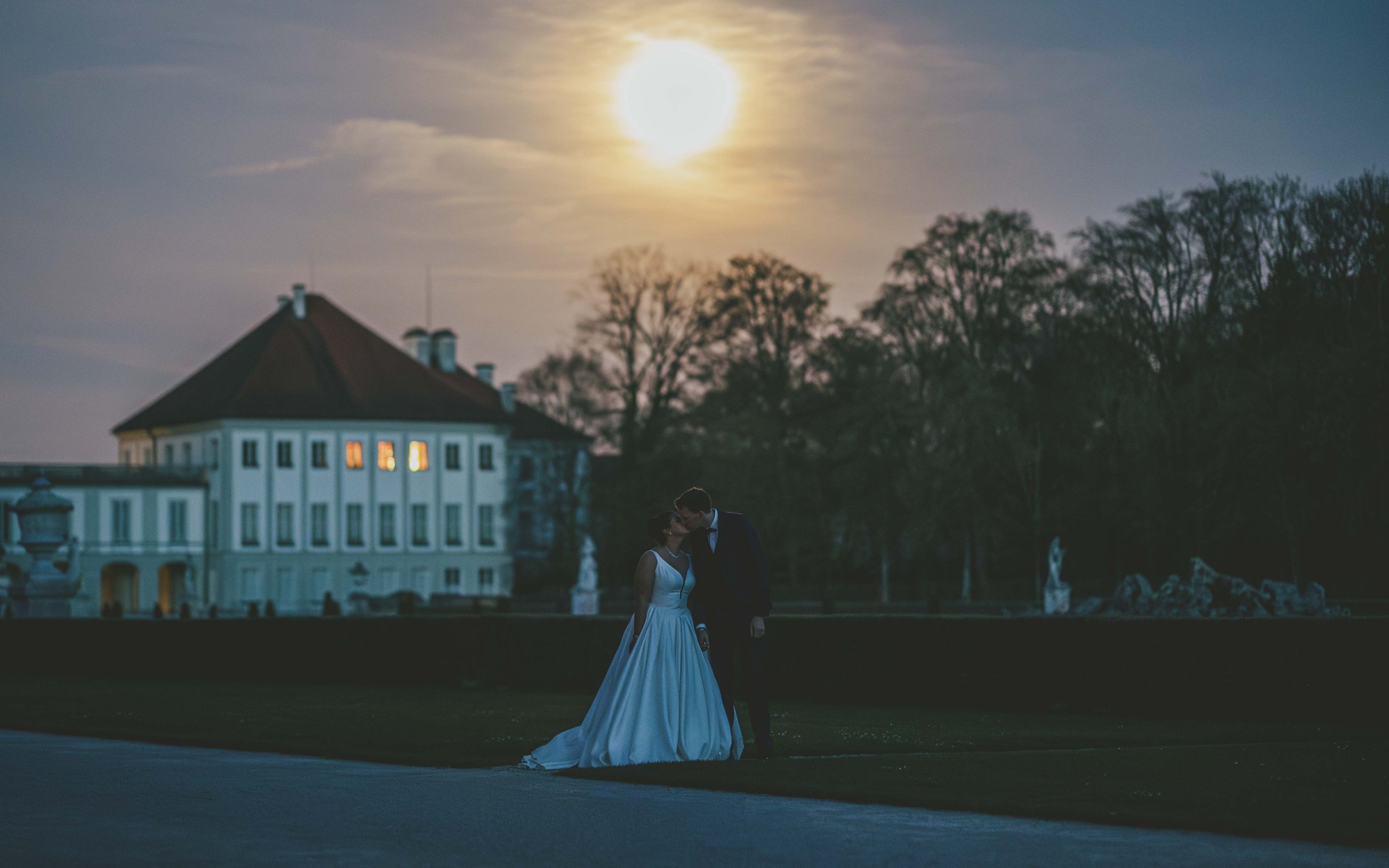 Ein Brautpaar küsst sich bei Sonnenuntergang vor einem großen, weißen Haus in einer parkähnlichen Umgebung.