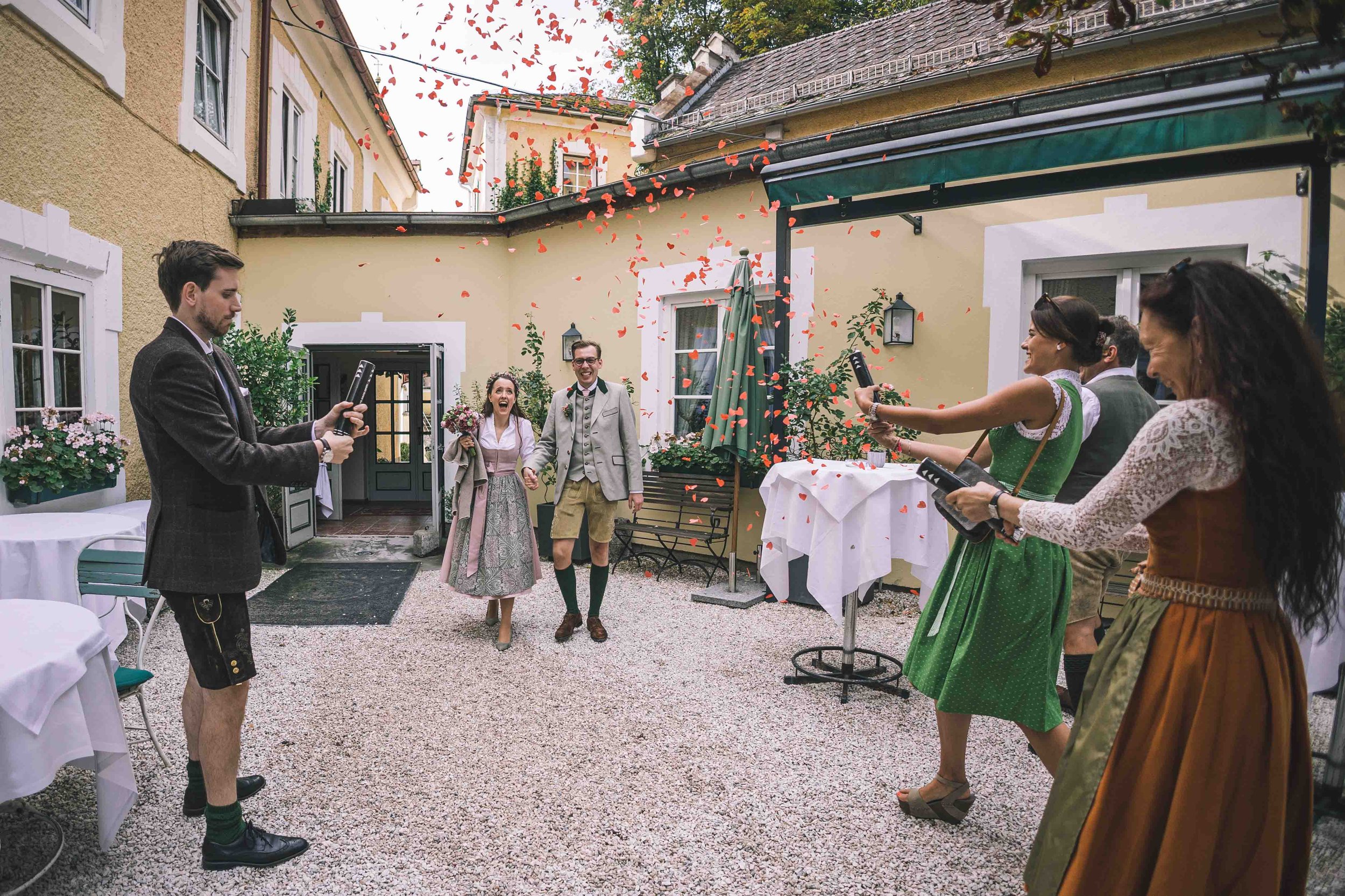 Hochzeitspaar läuft durch confettizerstreutes Gästepublikum im Außenhof eines Hauses in traditioneller bayrischer Tracht.