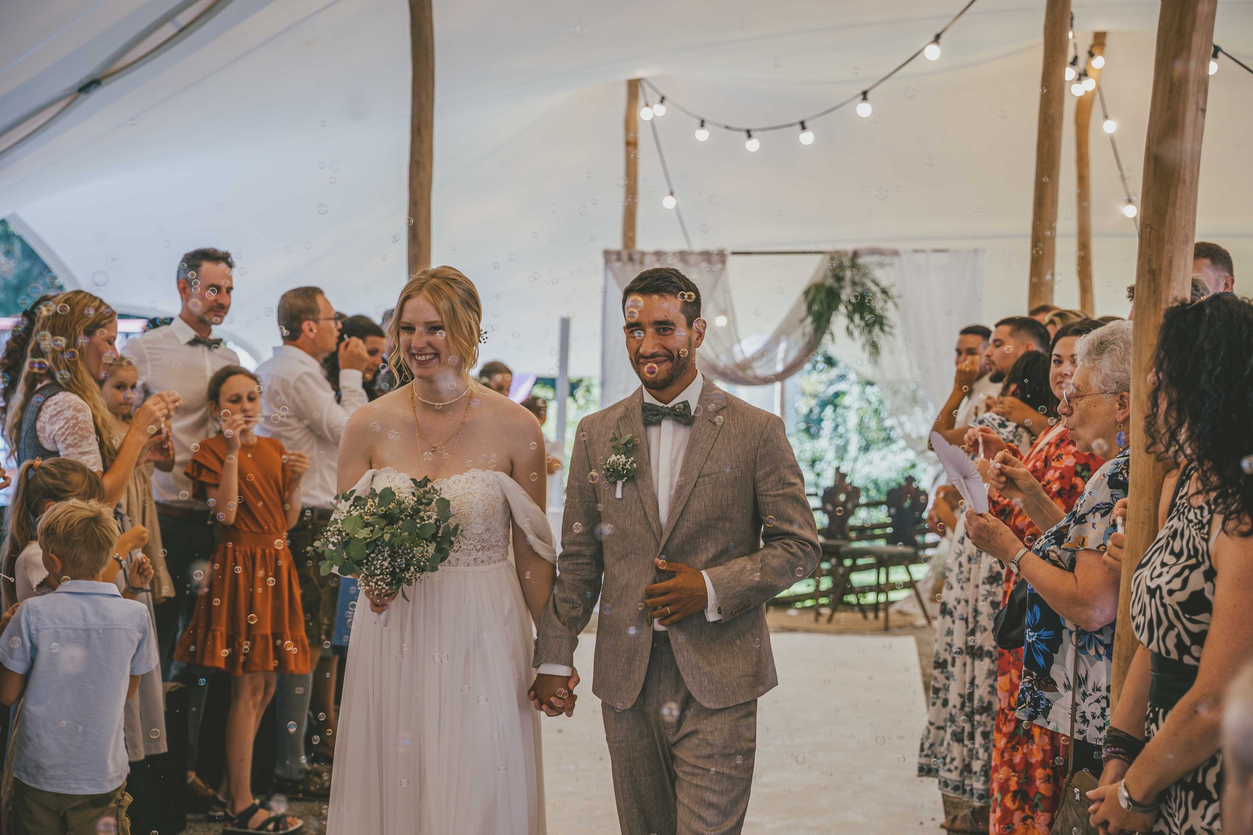 Ein Brautpaar hält Händchen bei ihrer Hochzeit, umgeben von Gästen, in einem festlich geschmückten Zelt mit Lichterketten.