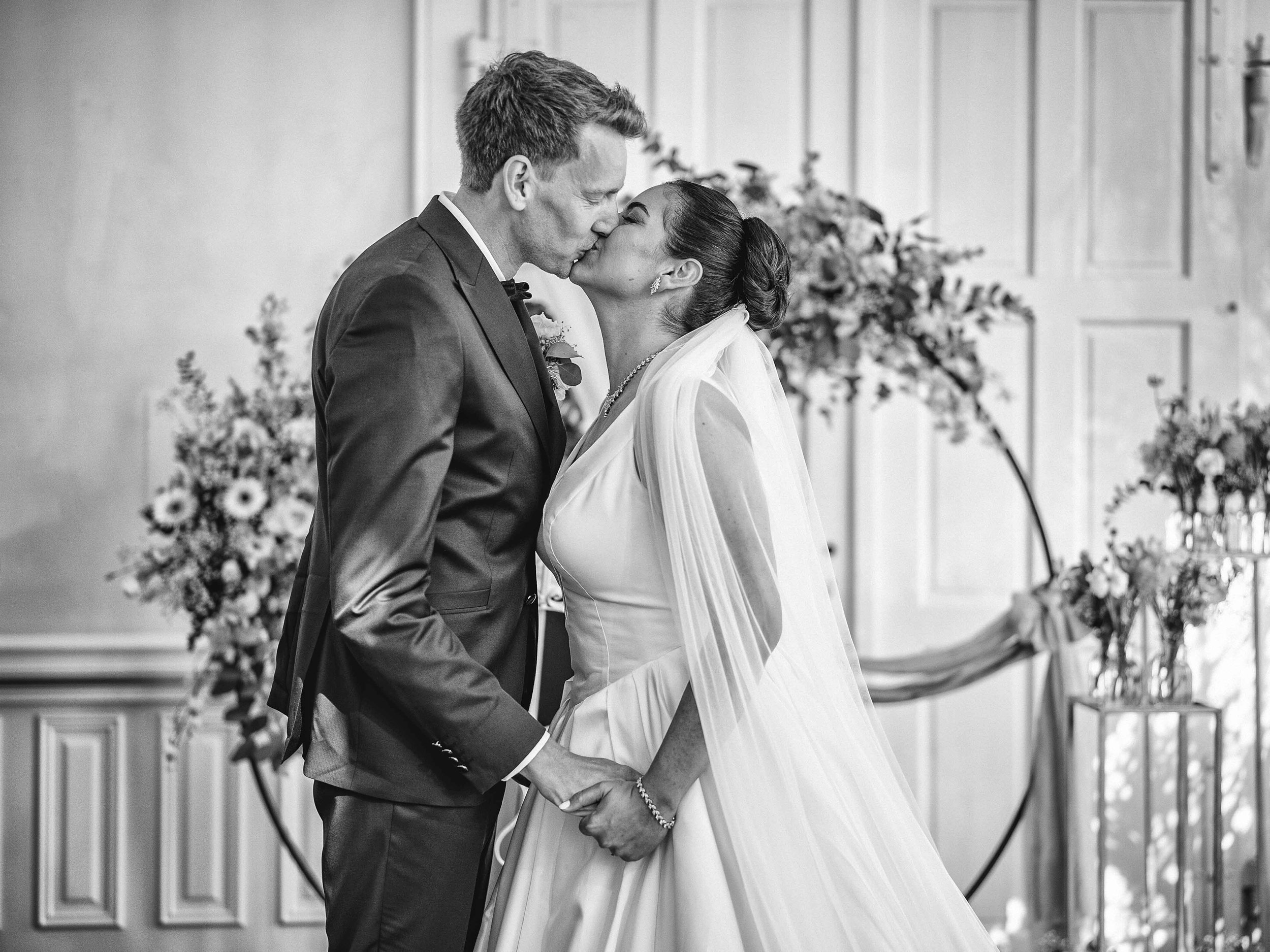 Ein Brautpaar küsst sich bei der Hochzeit, schwarz-weiß Foto, im Hintergrund Blumen und Dekoration.