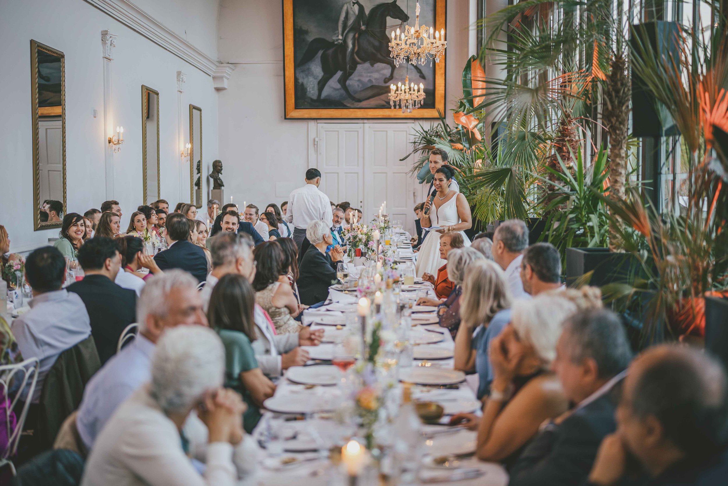Hochzeitstafel in eleganter Unterkunft mit vielen Gästen, eine Frau hält eine Rede, große Palme und Porträt an der Wand.