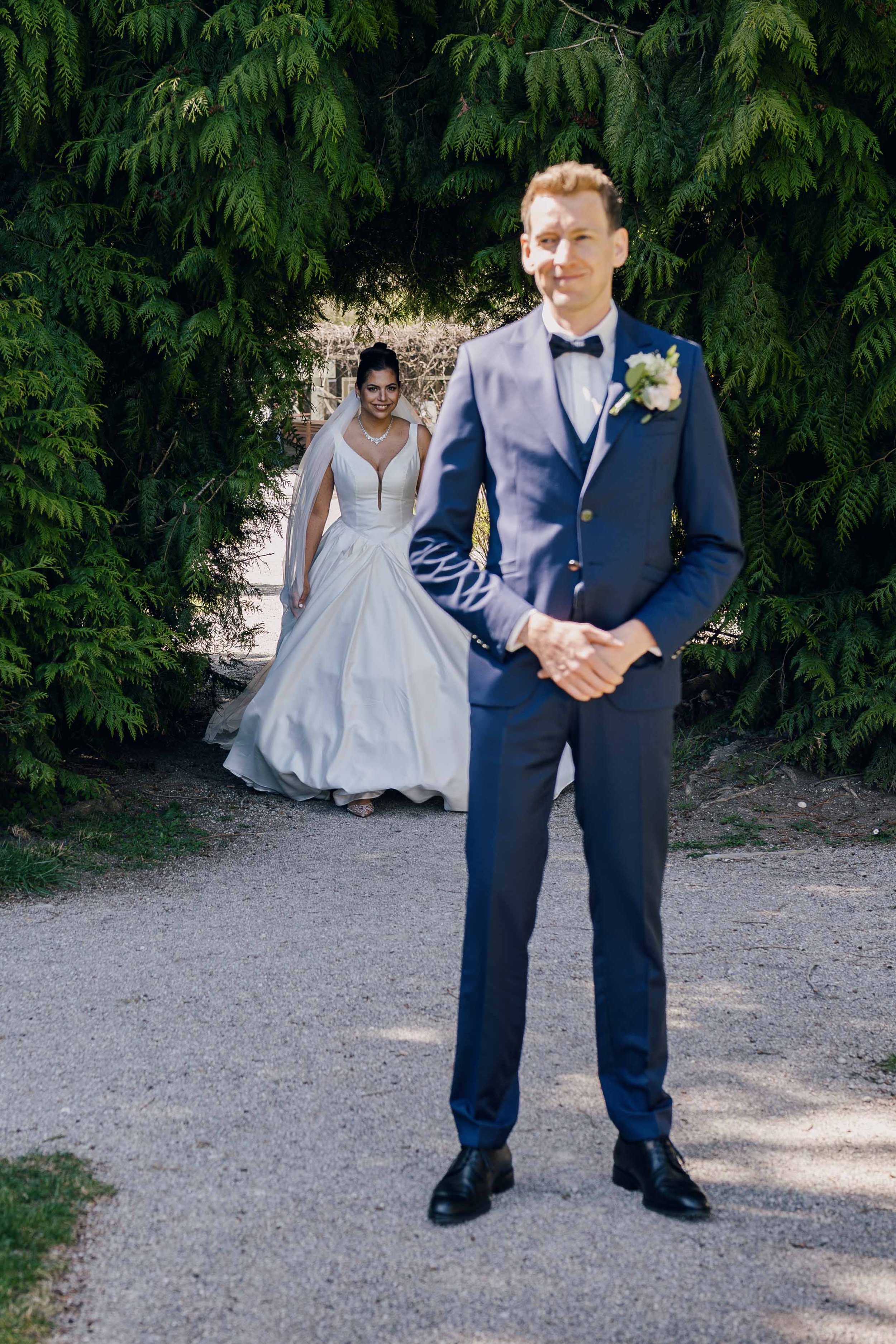 Ein Bräutigam steht im Vordergrund, trägt einen blauen Anzug mit Fliege und eine Blume am Revers, während eine lachende Braut im weißen Brautkleid im Hintergrund durch einen grünen Laubenbogen schaut.