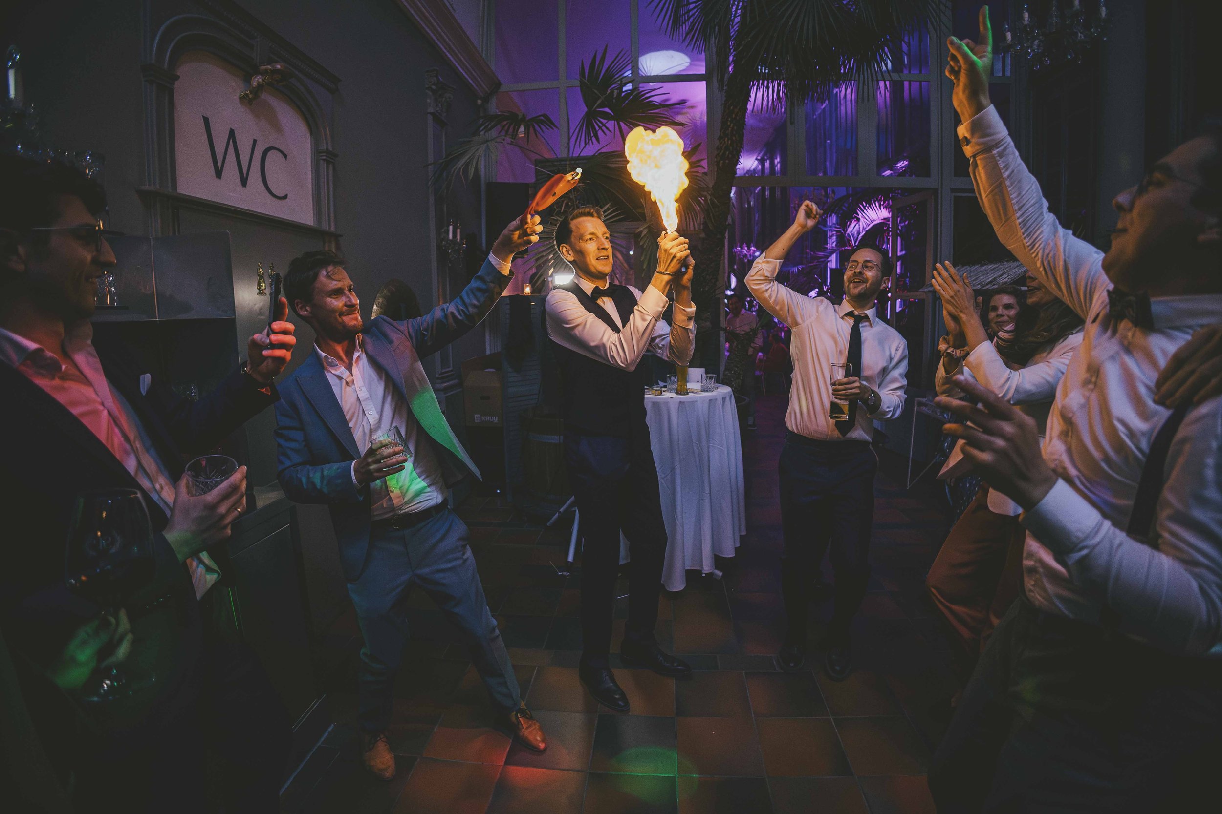Menschen feiern auf einer Party, einige mit Getränken, eine Person hält eine Flamme, alle scheinen Spaß zu haben.