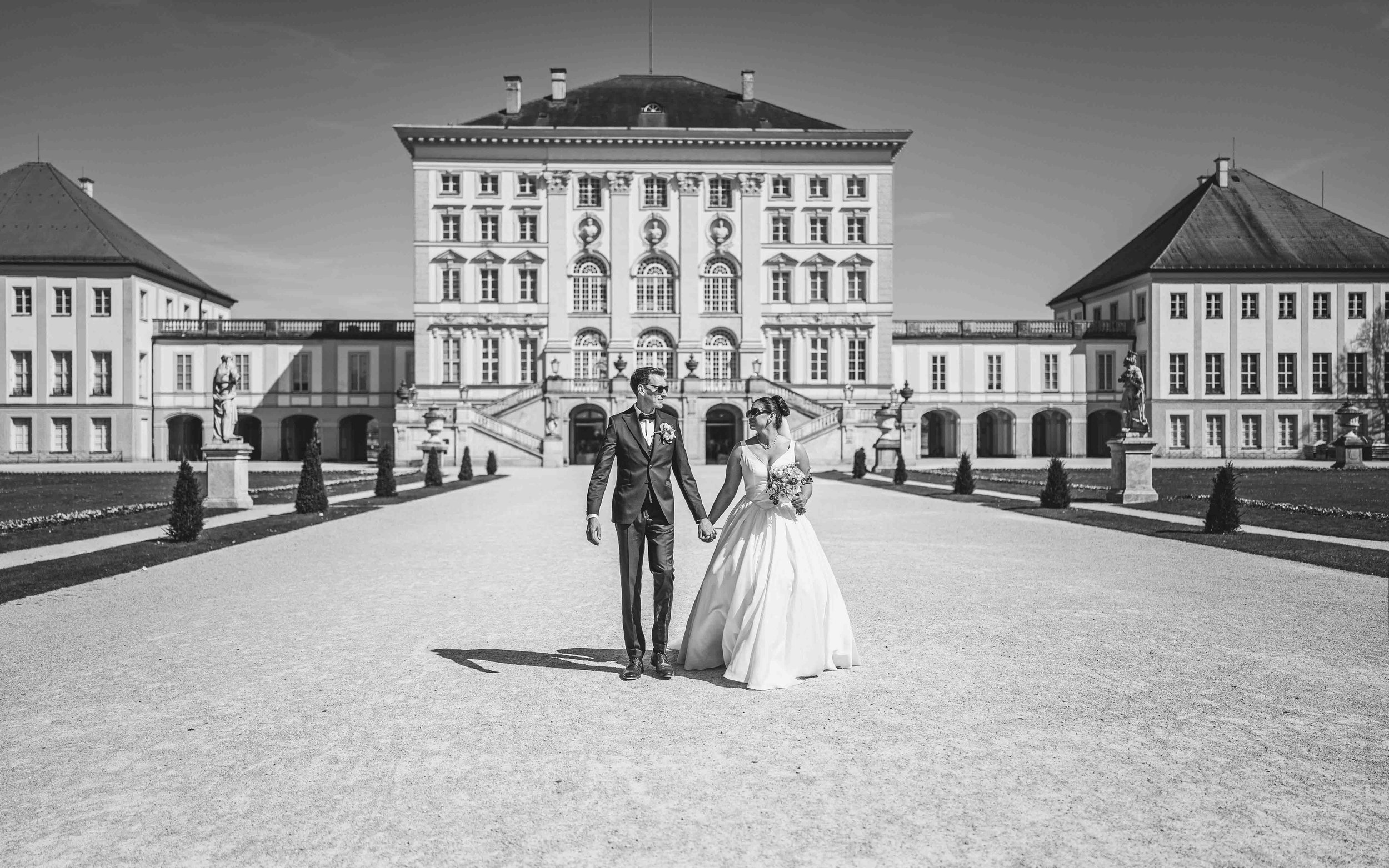 Ein frisch verheiratetes Paar in formeller Kleidung hält Händchen vor einem großen historischen Gebäude,Nümphenburg, München.