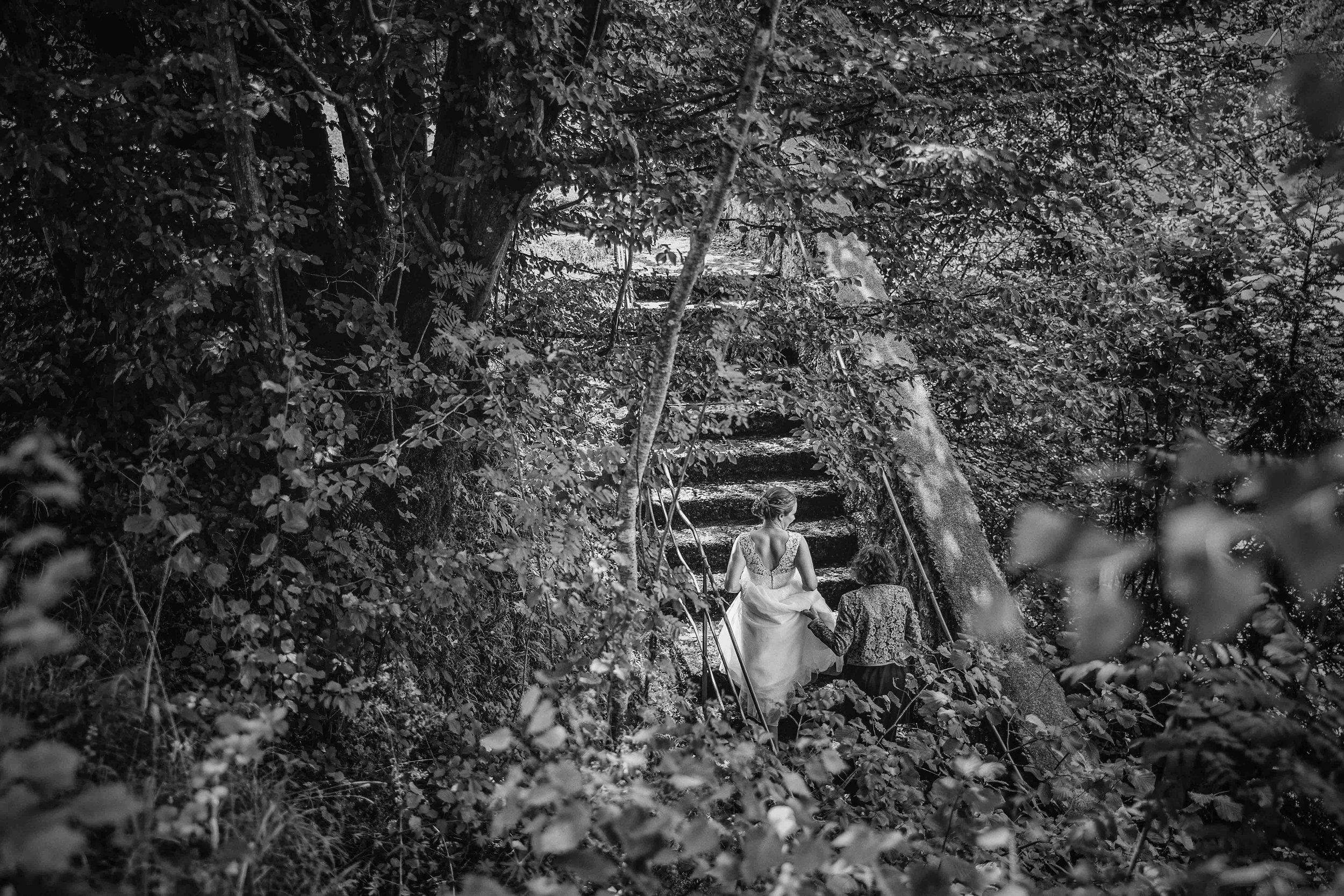 Schwarz-Weiß-Foto von einer Braut, die auf einer Waldtreppe steigt, begleitet von einer weiteren Frau, umgeben von Bäumen und dichten Blättern.