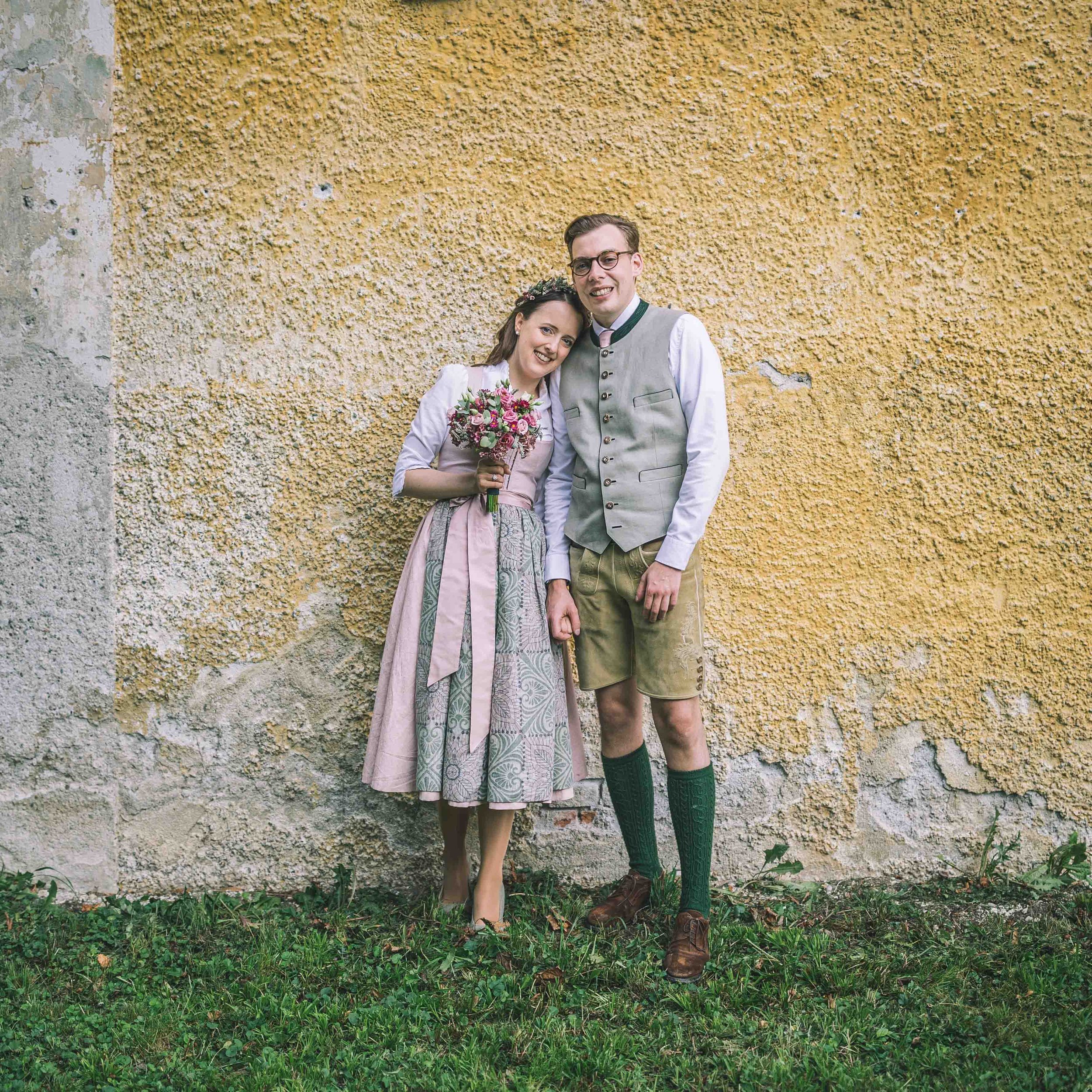 Ein ländliches Hochzeitsfoto von einem Paar in traditioneller bayerischer Kleidung, stehend vor einer gelben, textured Wand. Die Frau hält einen Blumenstrauß, beide lachen.