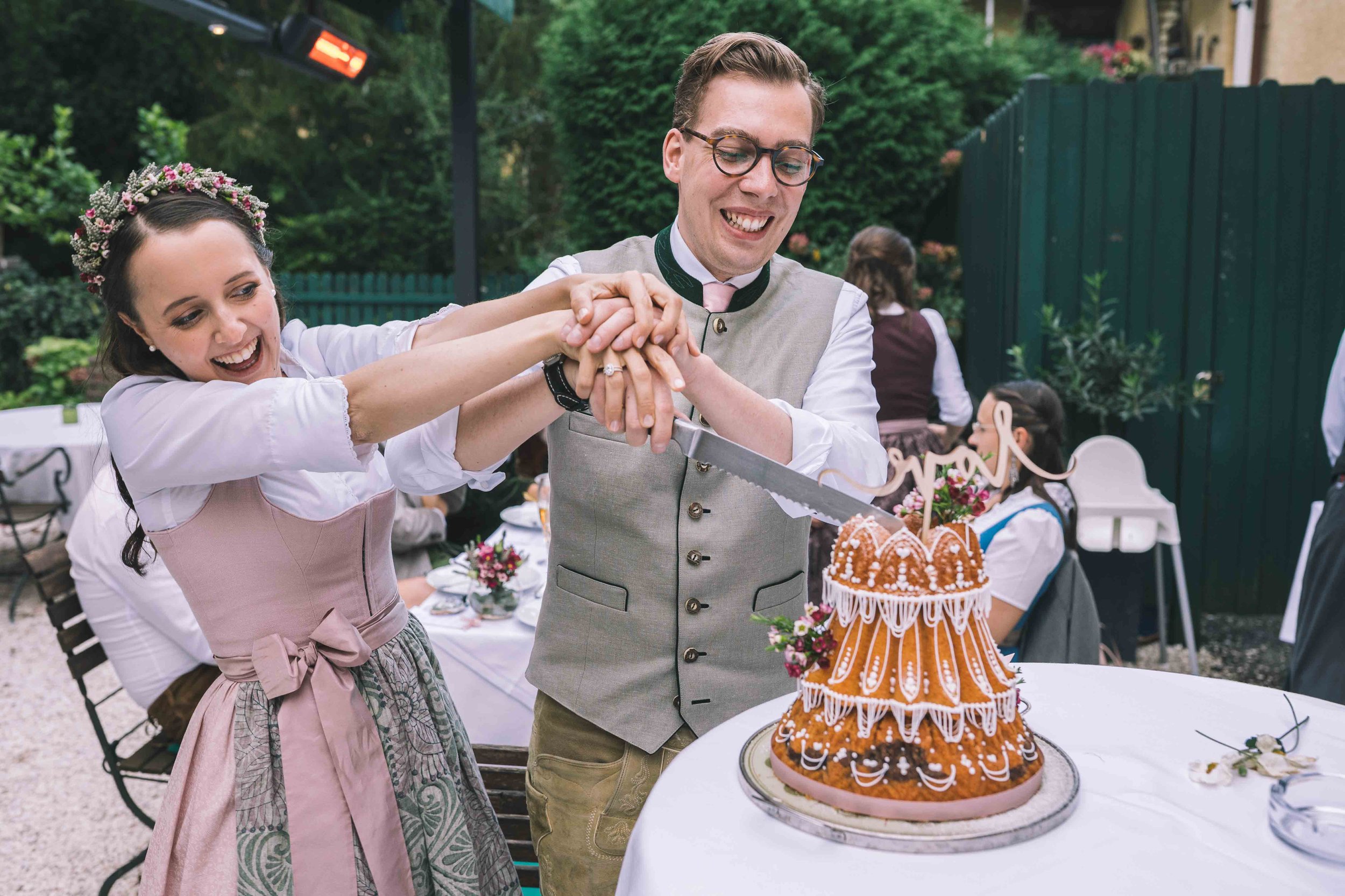 Ein lachendes Paar, das zusammen einen Hochzeitstorte anschneidet, bei einer Feier im Freien, umgeben von Gästen und Dekorationen.