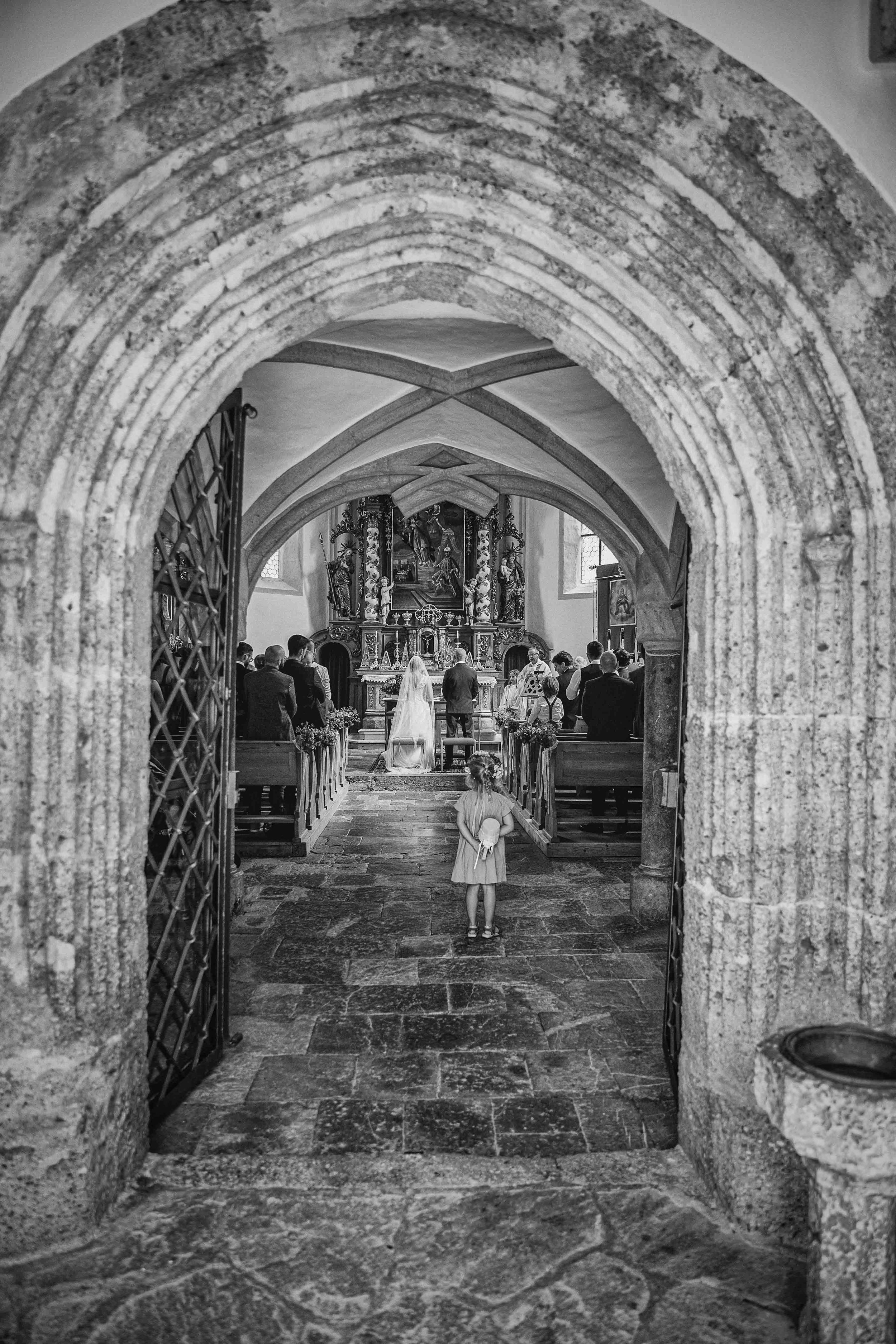 Schwarz-Weiß-Foto einer Hochzeit in einer Kirche, gesehen durch einen alten Steingarten. Im Hintergrund steht die Braut in weißem Kleid und Schleier an Altart, mit Gästen im Kirchenbankkreis. Im Vordergrund sitzt ein kleines Mädchen mit Blumen im Haa