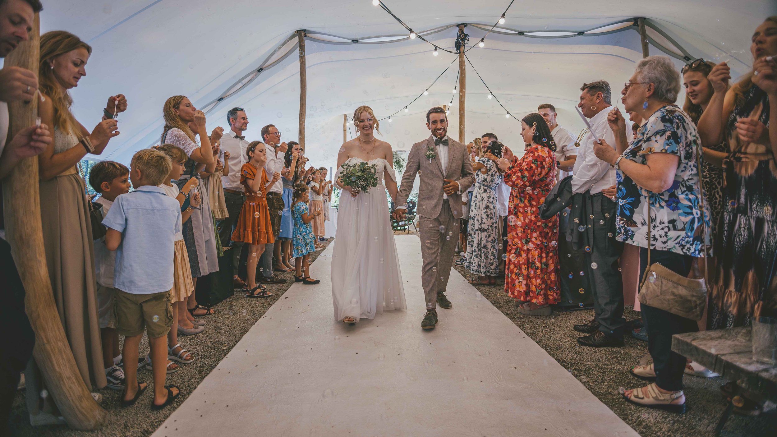 Ein Brautpaar ist in einem Zelt während einer Hochzeit, umgeben von lachenden Gästen, die Seifenblasen blasen.