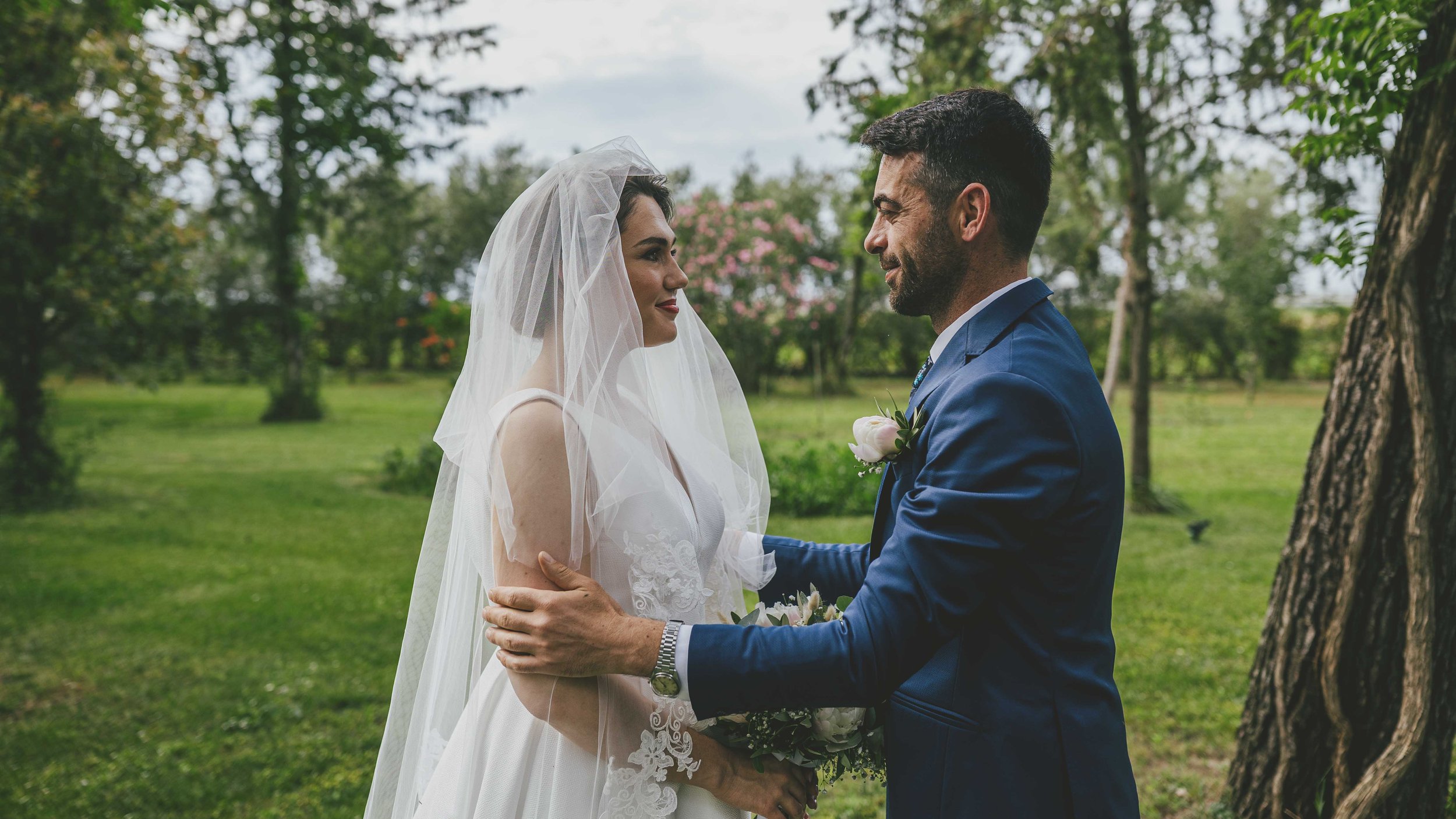 Ein Brautpaar steht im Park, die Frau trägt ein weißes Hochzeitskleid mit Schleier, der Mann trägt einen blauen Anzug mit Blumen im Revers, sie schauen sich liebevoll an.