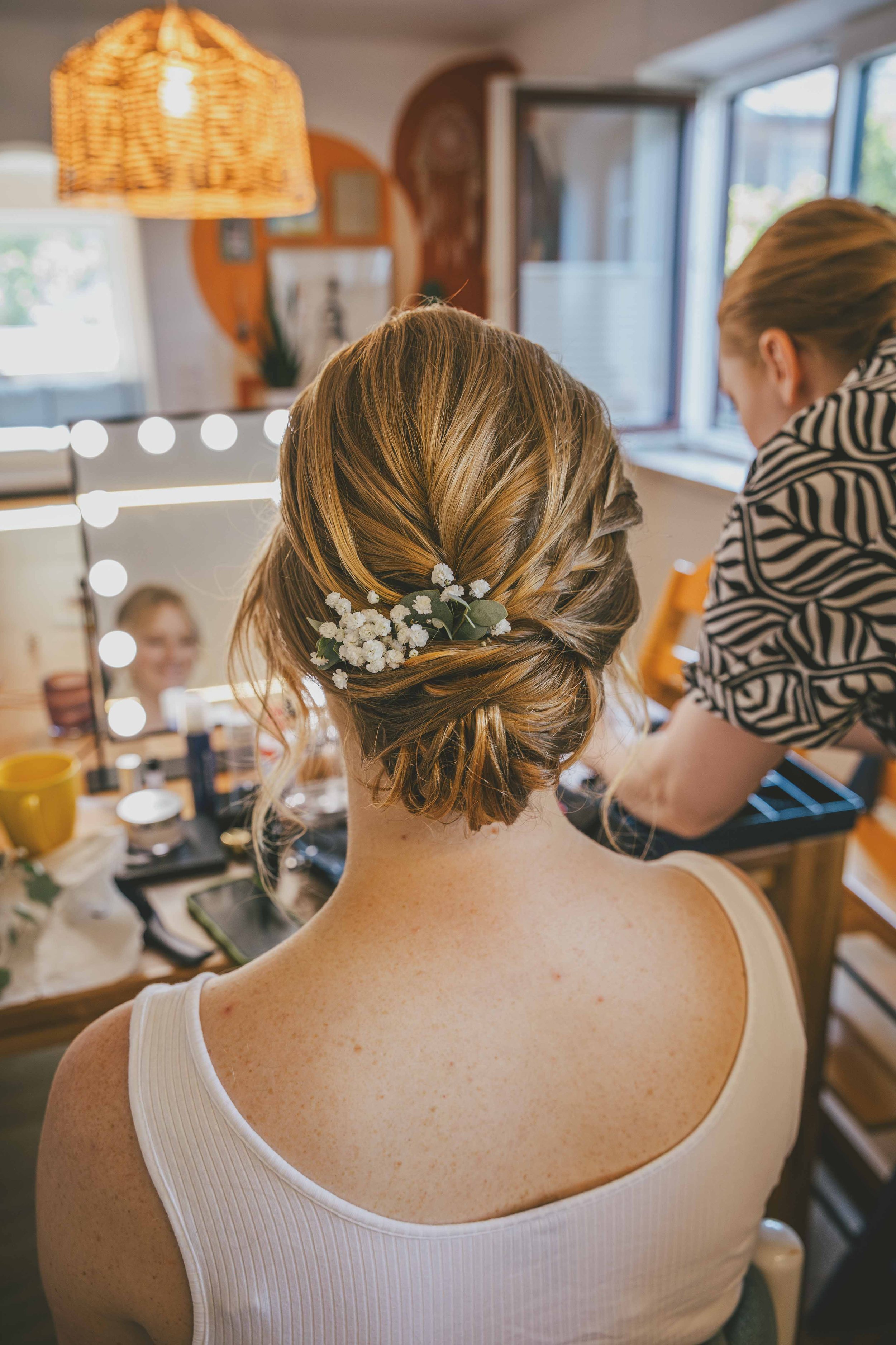 Frau mit Hochsteckfrisur und Blumenaccessoire, im Hintergrund wird Make-up für einen besonderen Anlass gemacht.