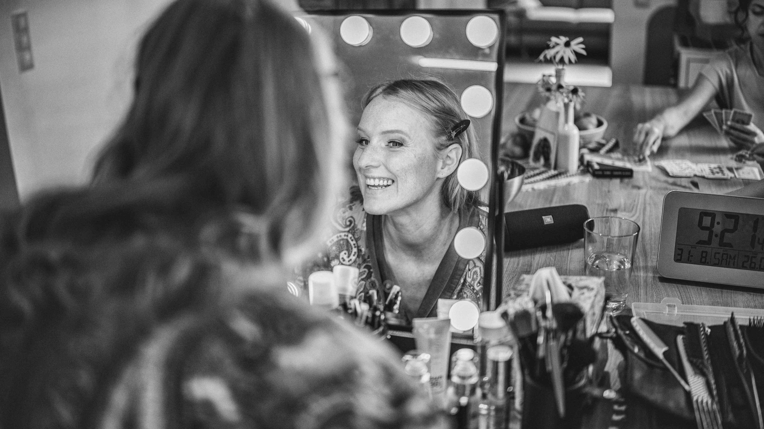 Eine Frau schaut in einen Spiegel mit Beleuchtung, während sie Make-up macht. Im Hintergrund sind weitere Personen, eine digitale Uhr und ein Tisch mit verschiedenen Gegenständen zu sehen.