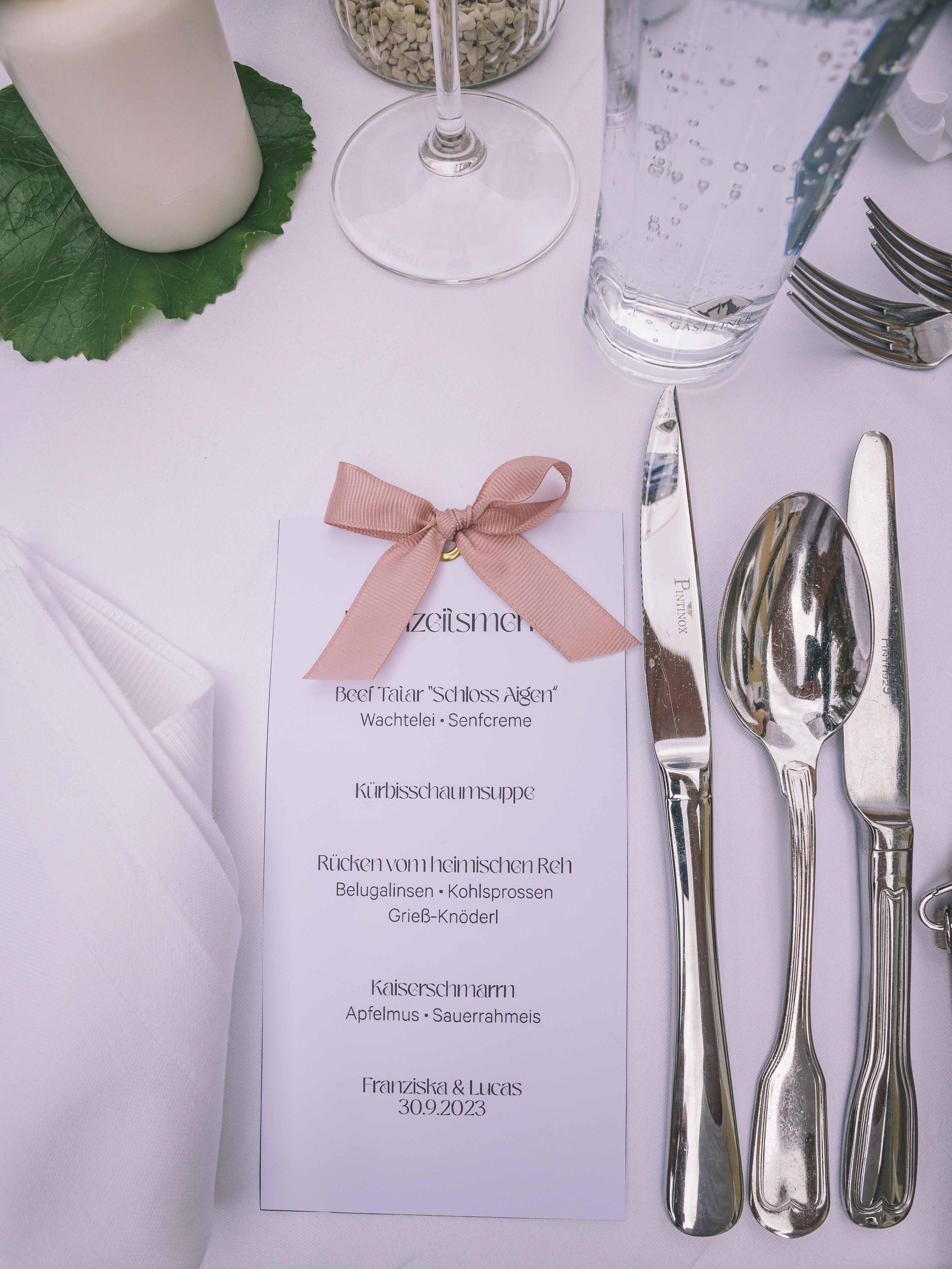 Menükarte auf einem Festtafel, dekoriert mit einem rosa Band, mittig neben Besteck und Wasser- und Weinglas, mit weiteren Besteckteilen und einer weißen Stoffserviette am Rand.