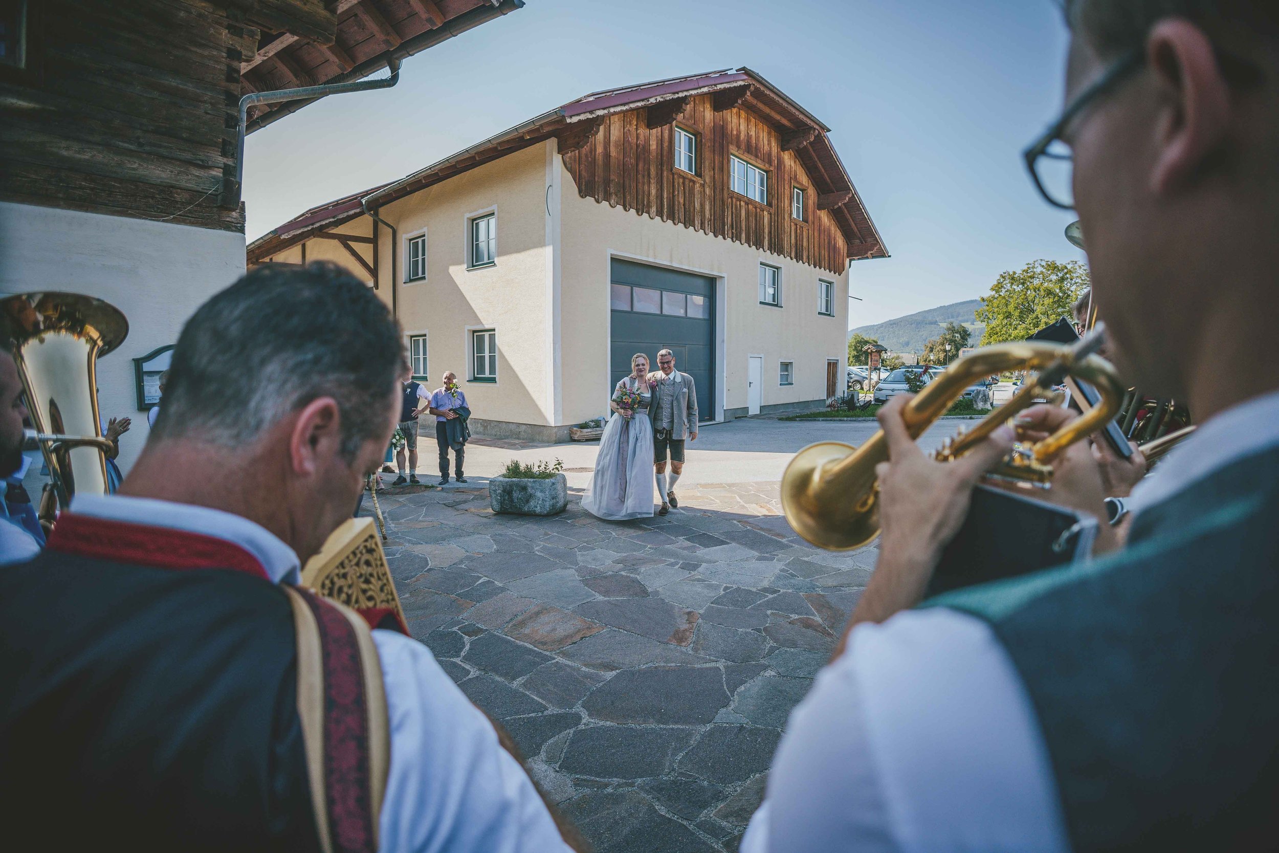 Braut und Bräutigam beim Spalier stehen vor einem Haus, während eine Musikkapelle im Vordergrund spielt.