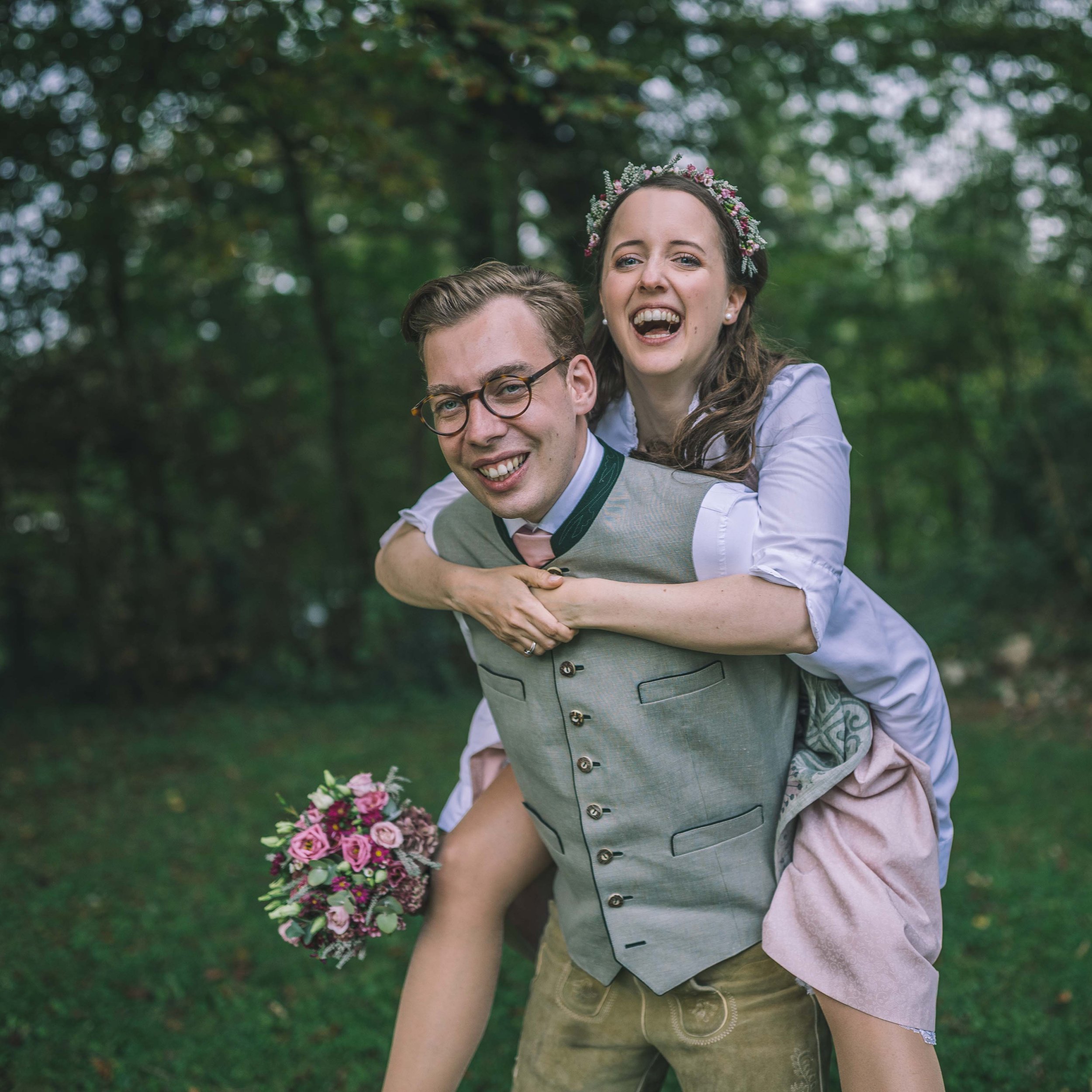 Ein lachendes Paar bei einer Hochzeit im Freien, der Mann trägt eine grau Weste und eine Brille, die Frau trägt ein weißes Kleid mit Blumenschmuck im Haar und hält einen Blumenstrauß.