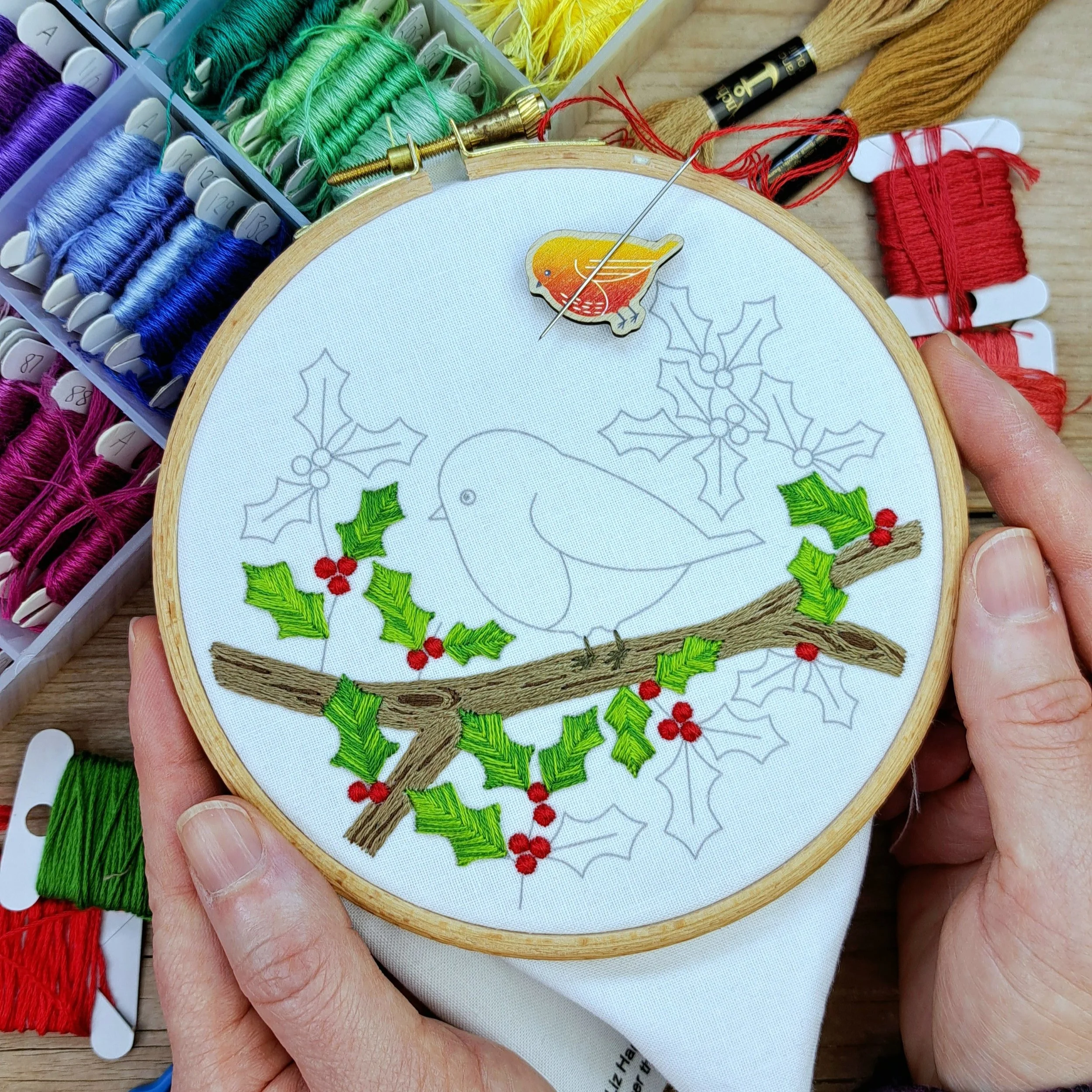 Partly completed robin on a holly branch, with branch, a few leaves, berries, and the robin’s feet stitched.