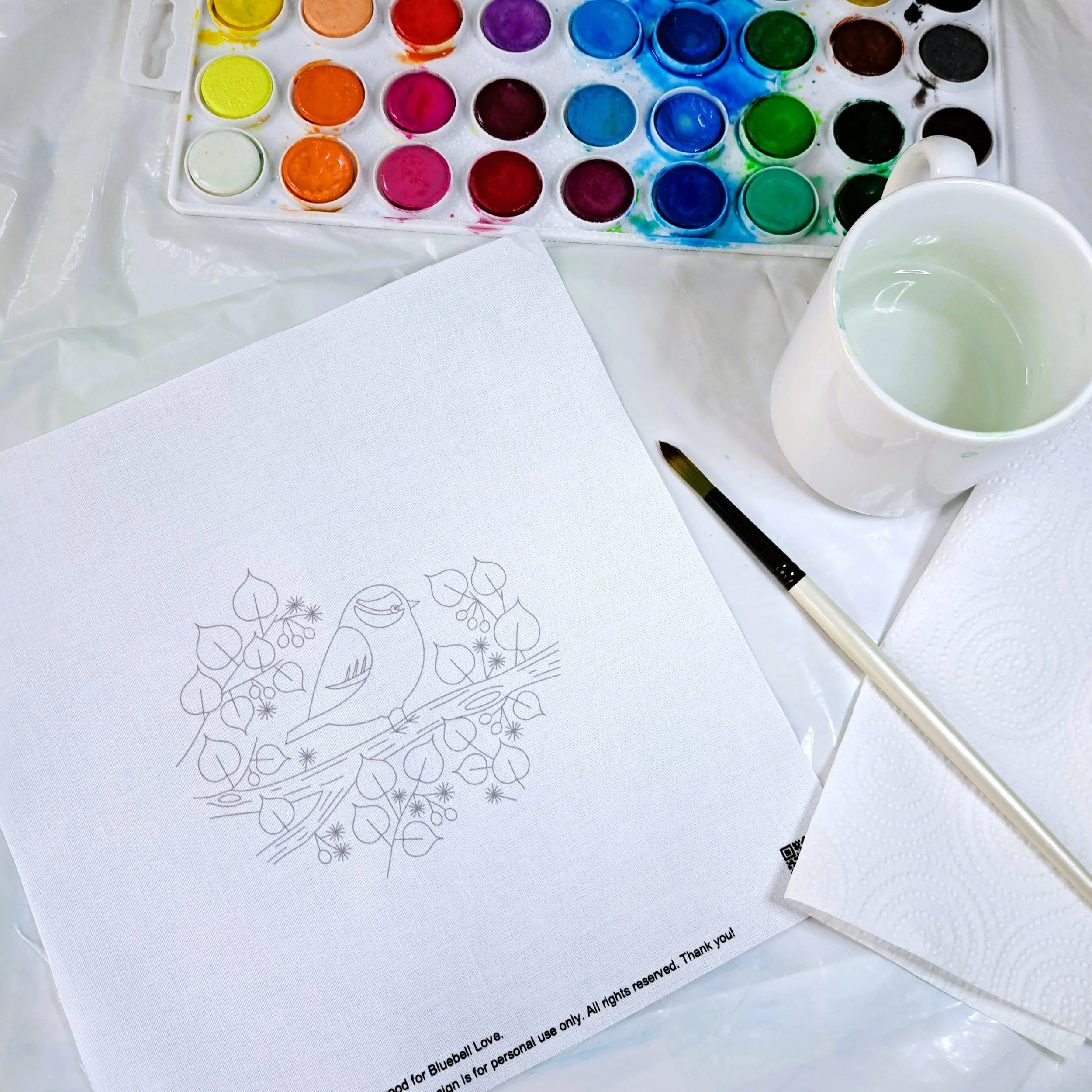 The set up for adding the watercolour paints to the embroidery panel. The fabric panel is laying on a plastic sheet, and the watercolour paints, a cup of water, and paint brush are ready to use.