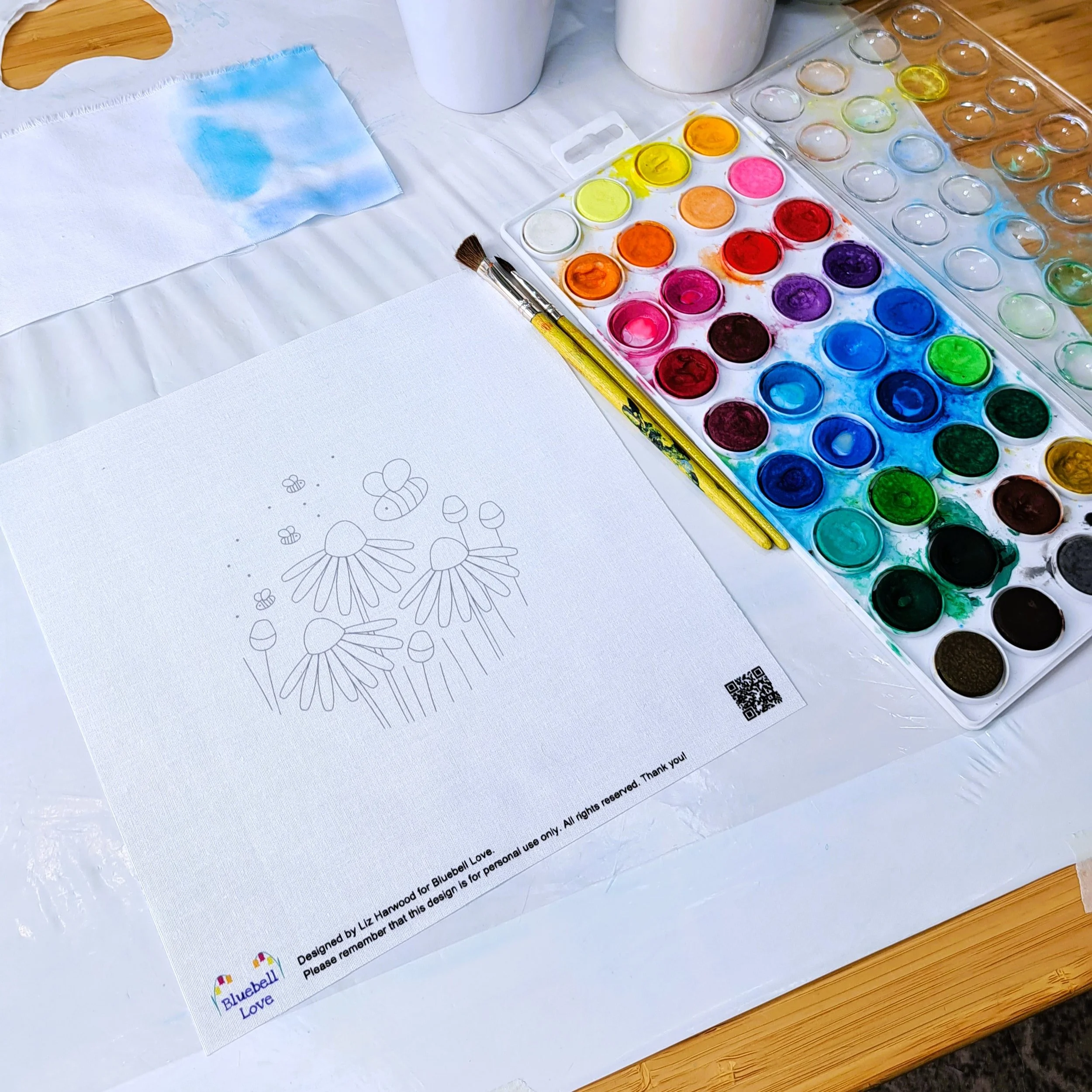 The set up for adding the watercolour paints to the embroidery panel. The fabric panel is laying on a plastic sheet, and the watercolour paints, a cup of water, and paint brush are ready to use.