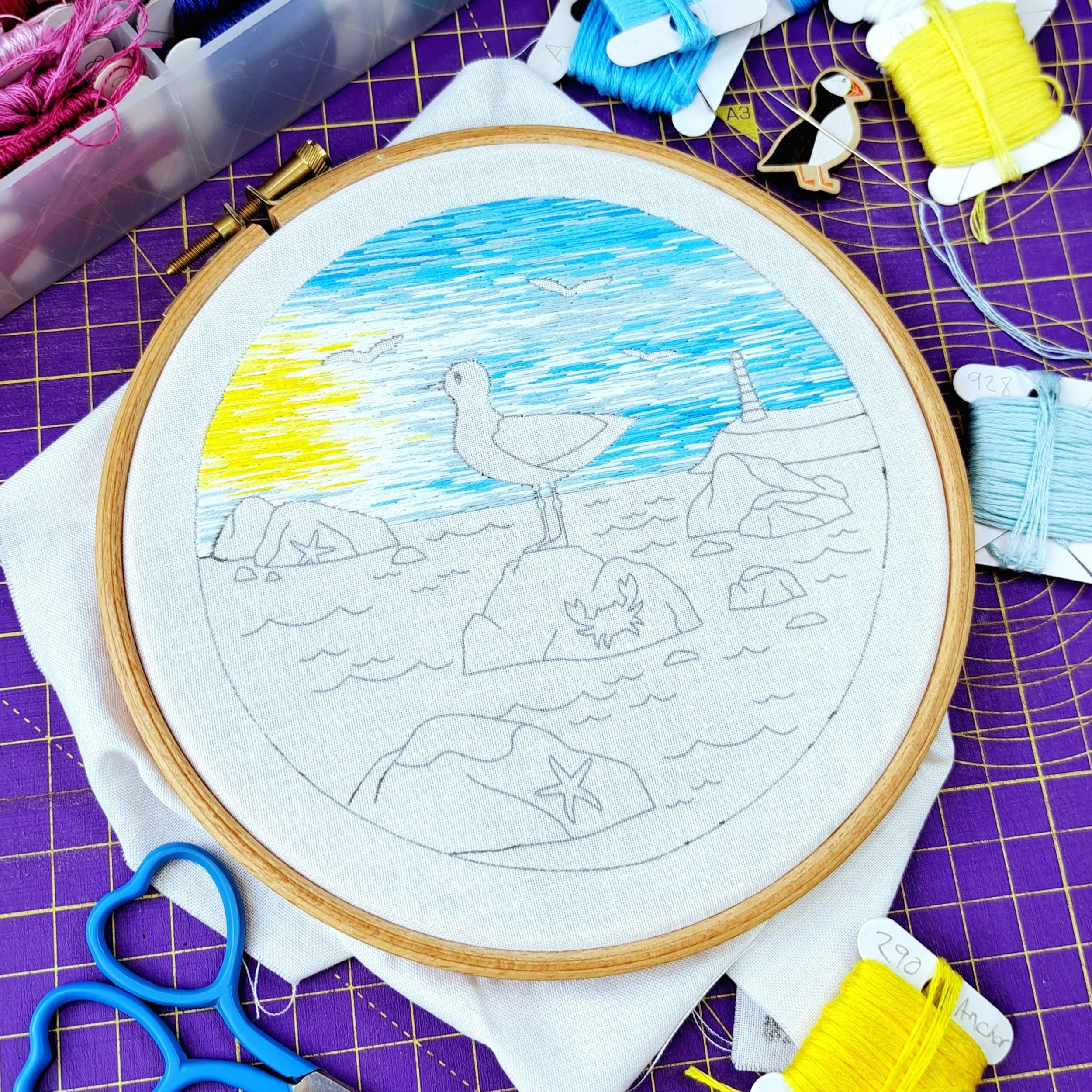 Partially stitched seagull embroidery panel in a 6-inch hoop on a studio desk surrounded by sewing tools
