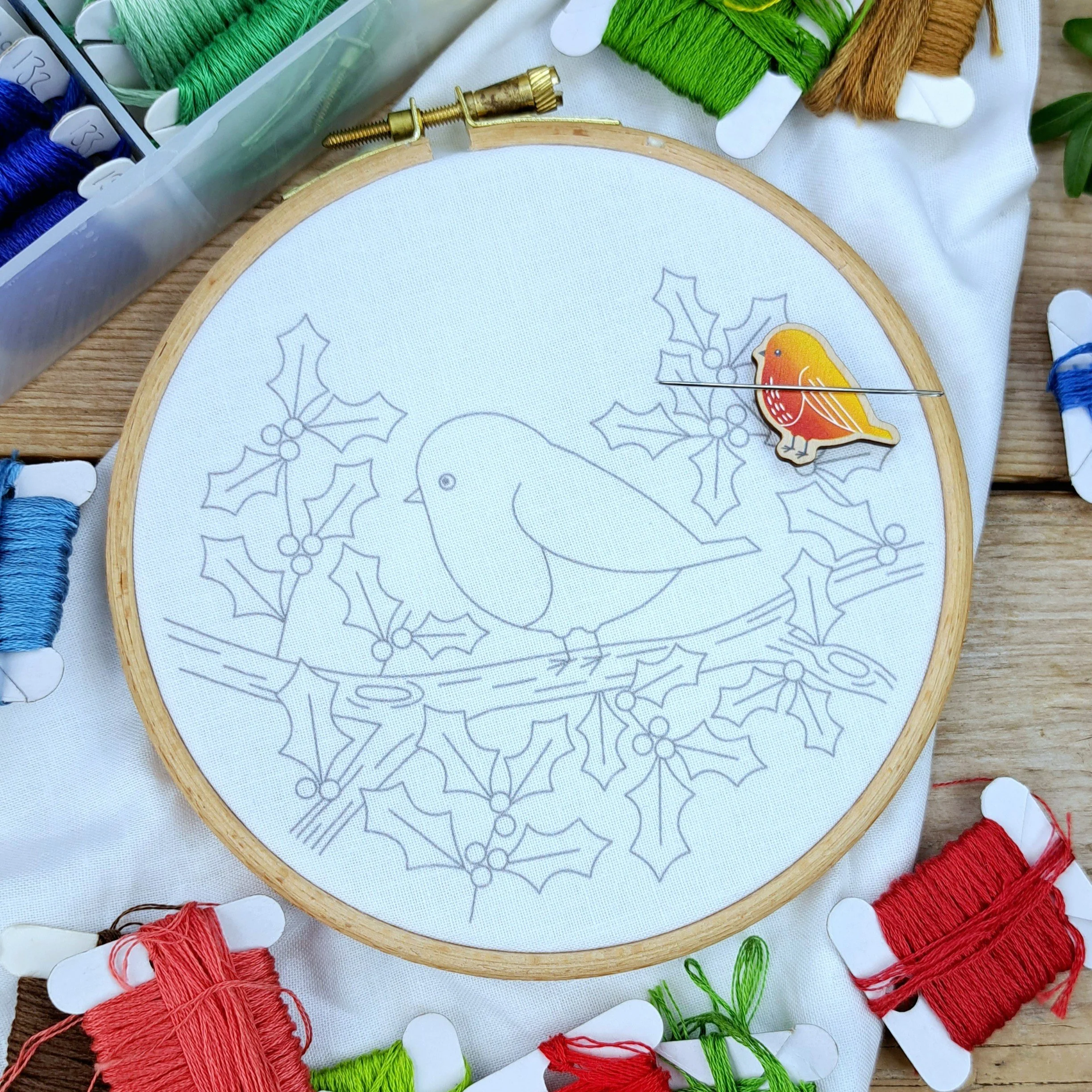 Unstitched robin embroidery panel in a hoop, ready for stitching, showing the bird on a holly branch.