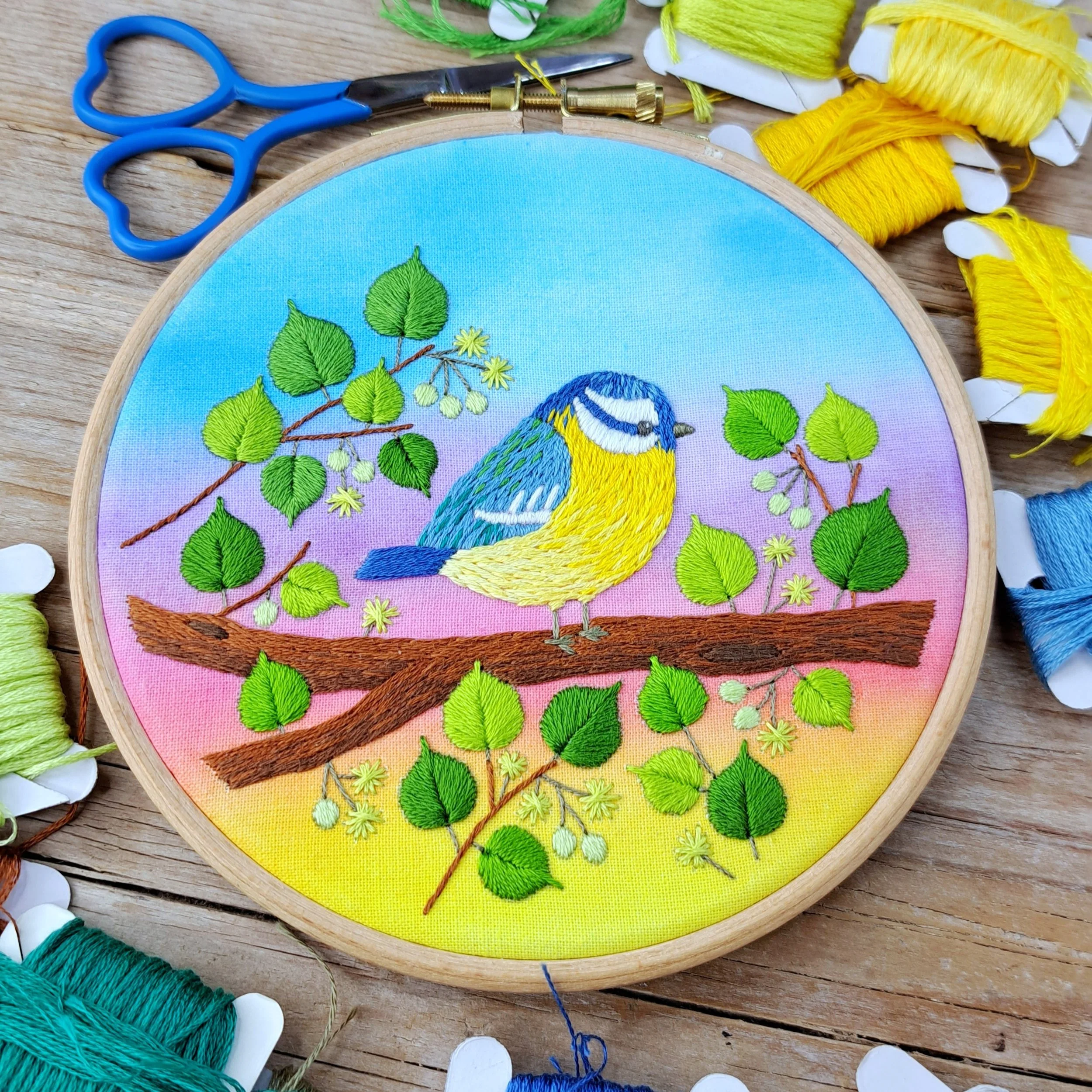 Finished embroidery of a colourful bird on a tree branch with a pretty watercolour painted sunrise background, displayed in an embroidery hoop.