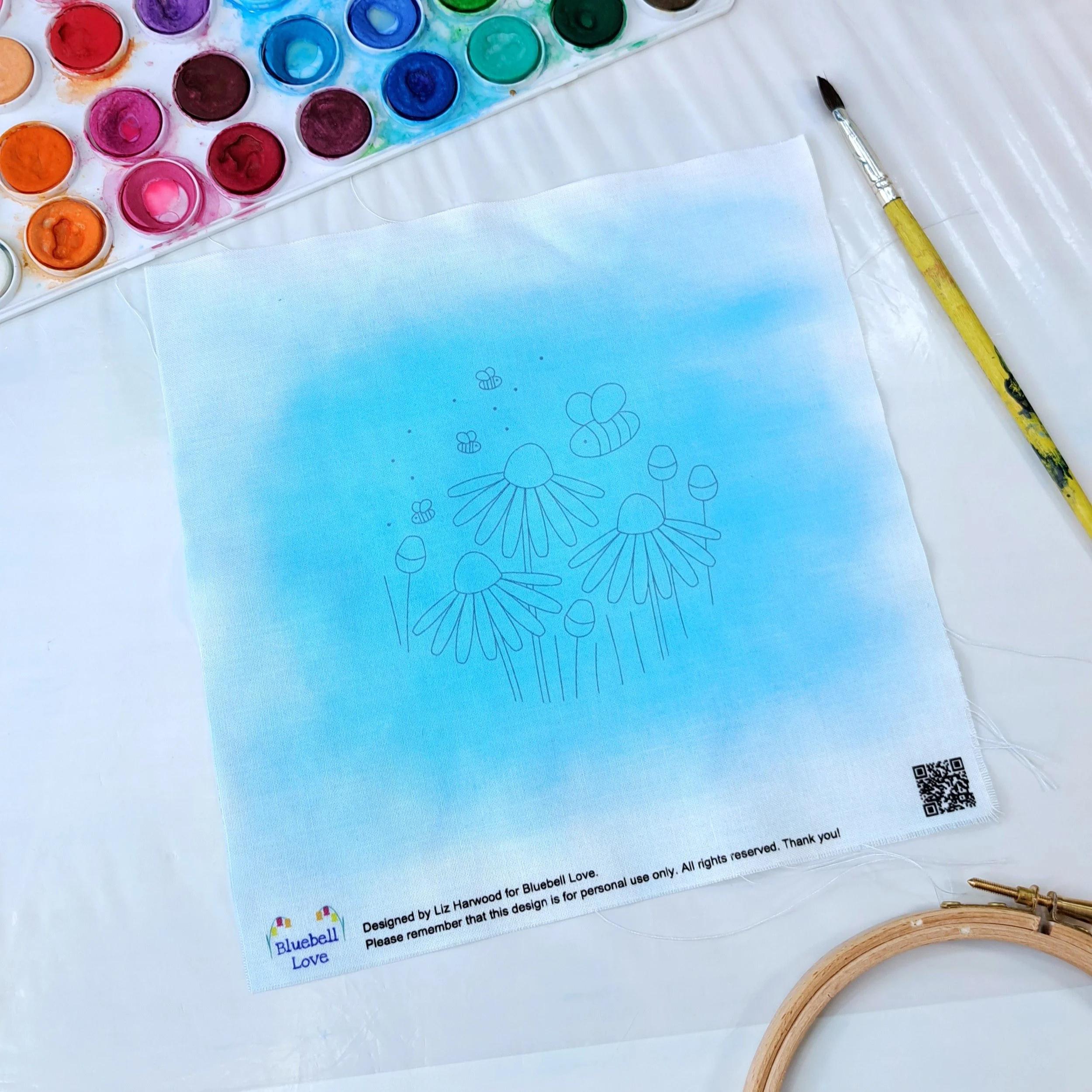 The fully painted flower and bee embroidery panel laid out flat, showing the completed watercolour blue background before stitching begins.