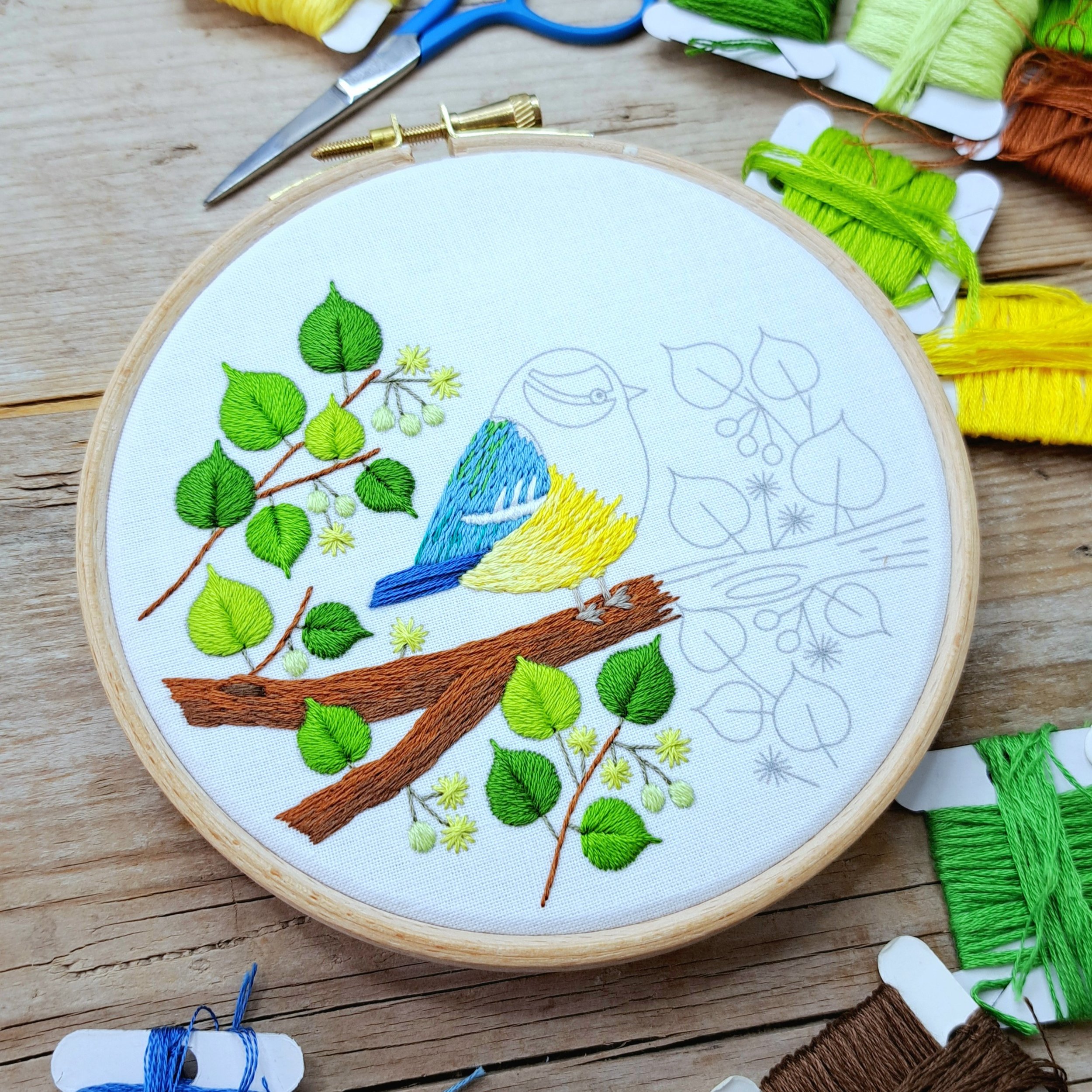 Work-in-progress blue tit bird embroidery panel in a 5-inch hoop, with colourful green leaves and blue and yellow feathers.