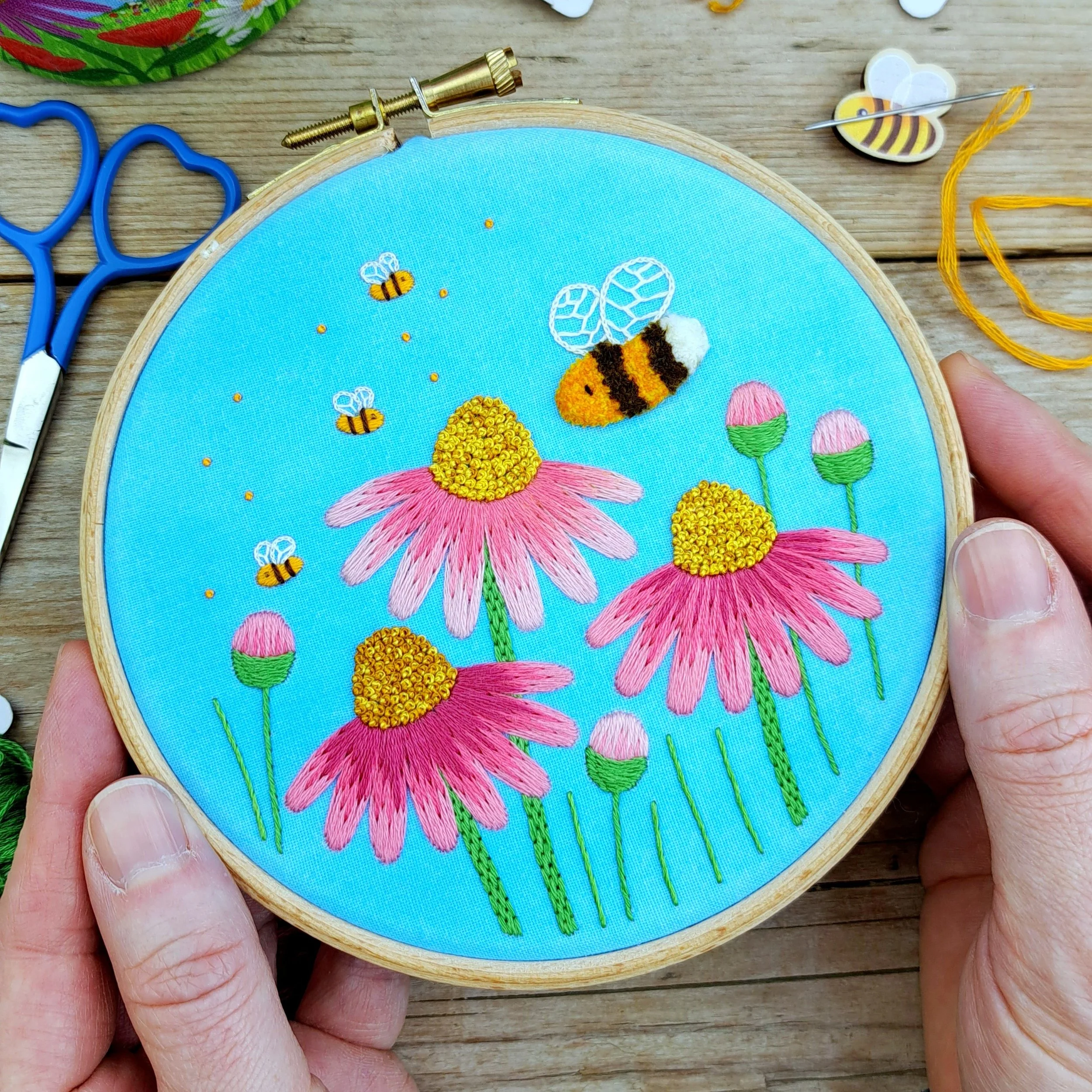 Finished embroidery of pink flowers, buds and flying bees with a blue watercolour painted background, displayed in an embroidery hoop and held in my hands.