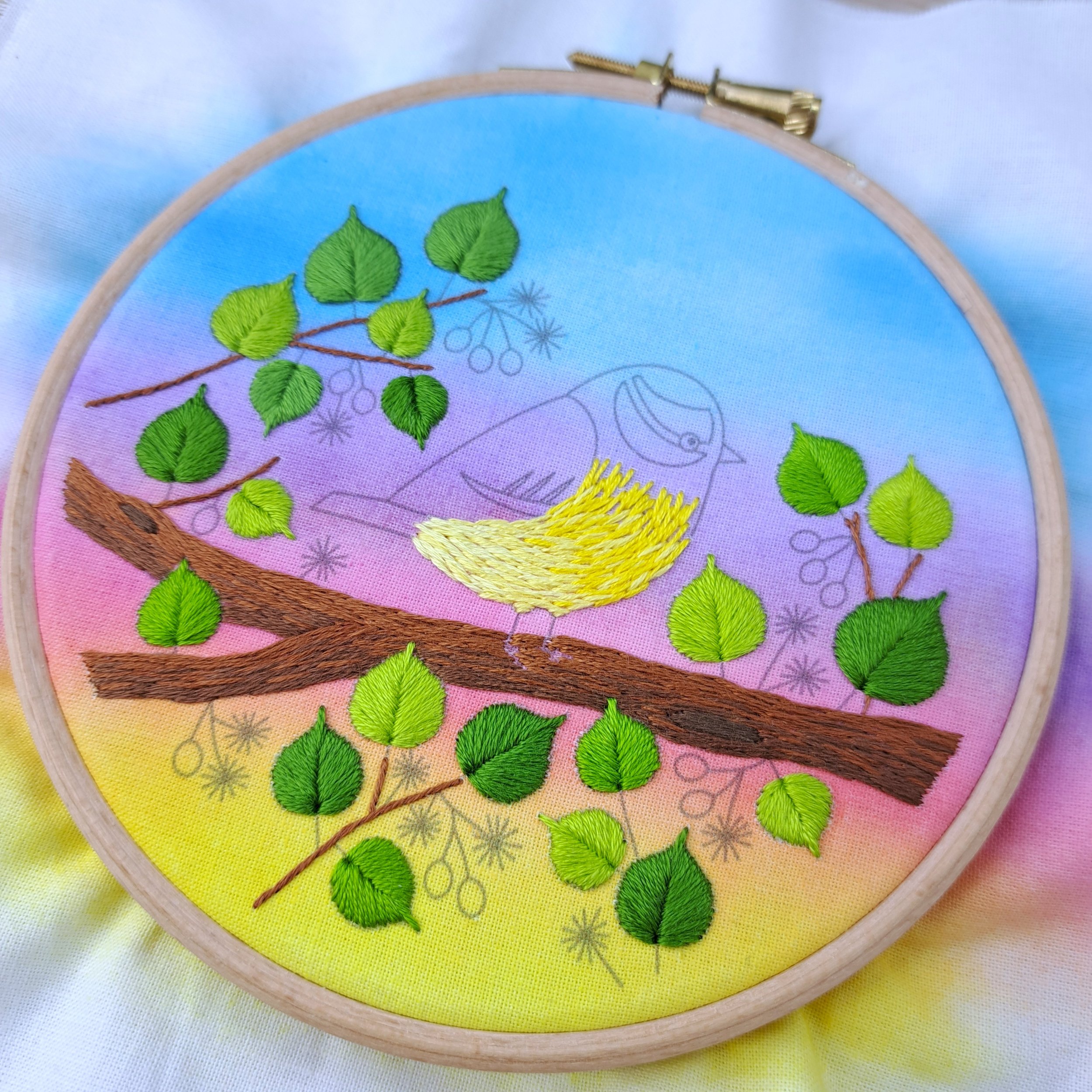 Partly completed embroidery of a bird on a branch embroidery design. The tree branch, the leaves, the twigs and the chest of the bird have been stitched.
