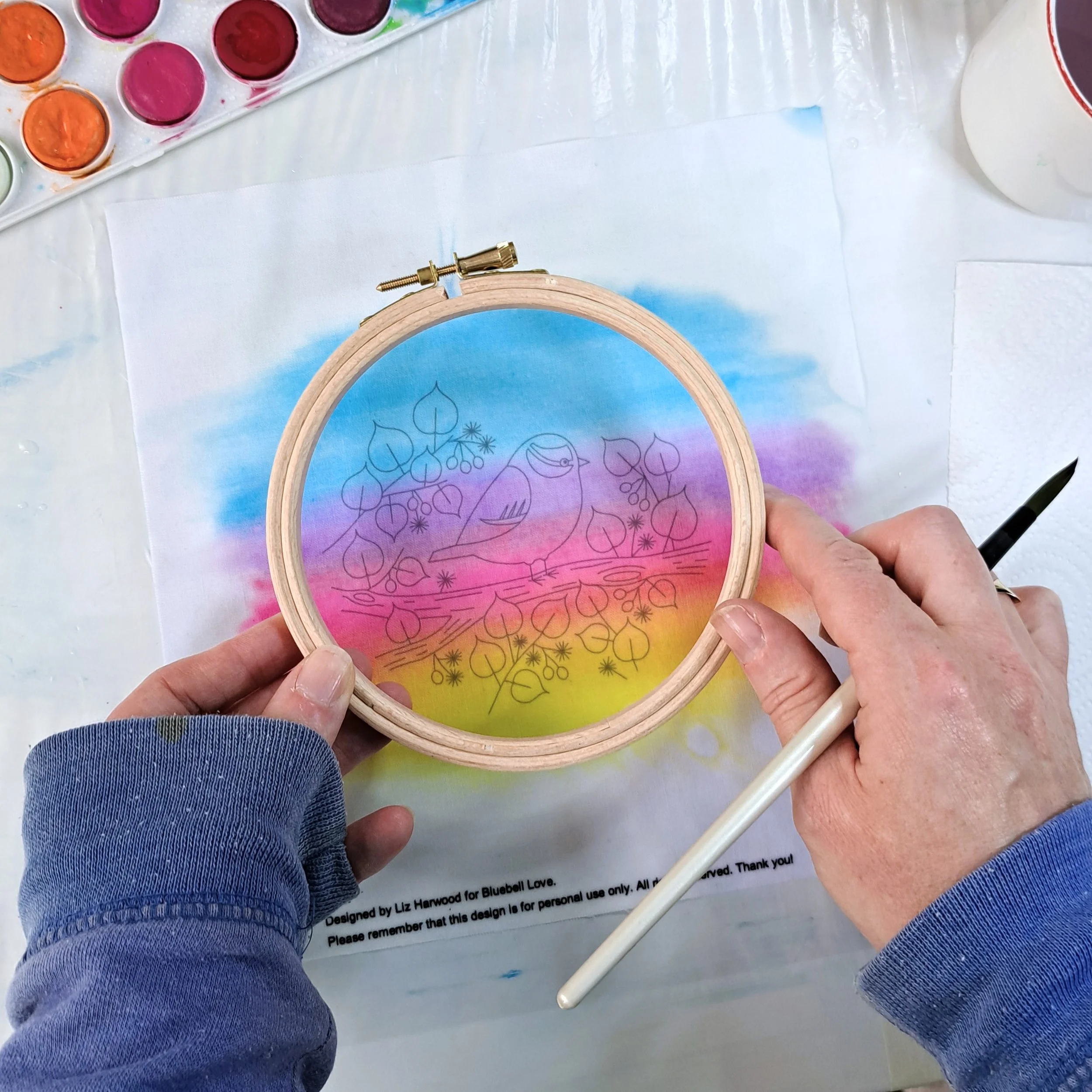 My hand painting holding an embroidery hoop over the painted fabric panel to make sure I’ve painted a little beyond the edges of the wooden hoop.