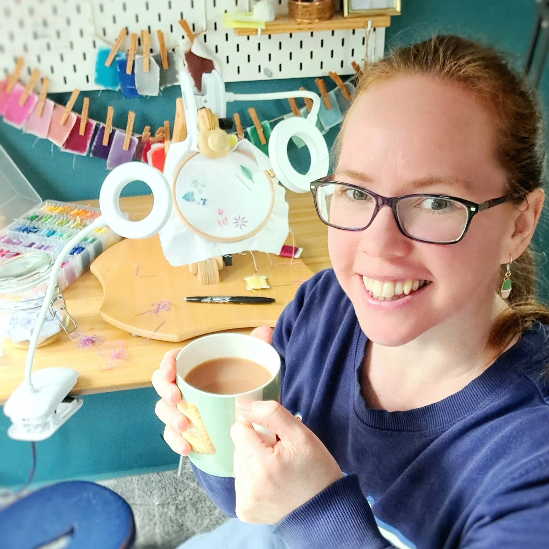 Liz holding a cup of tea in her embroidery studio with stitching supplies behind her