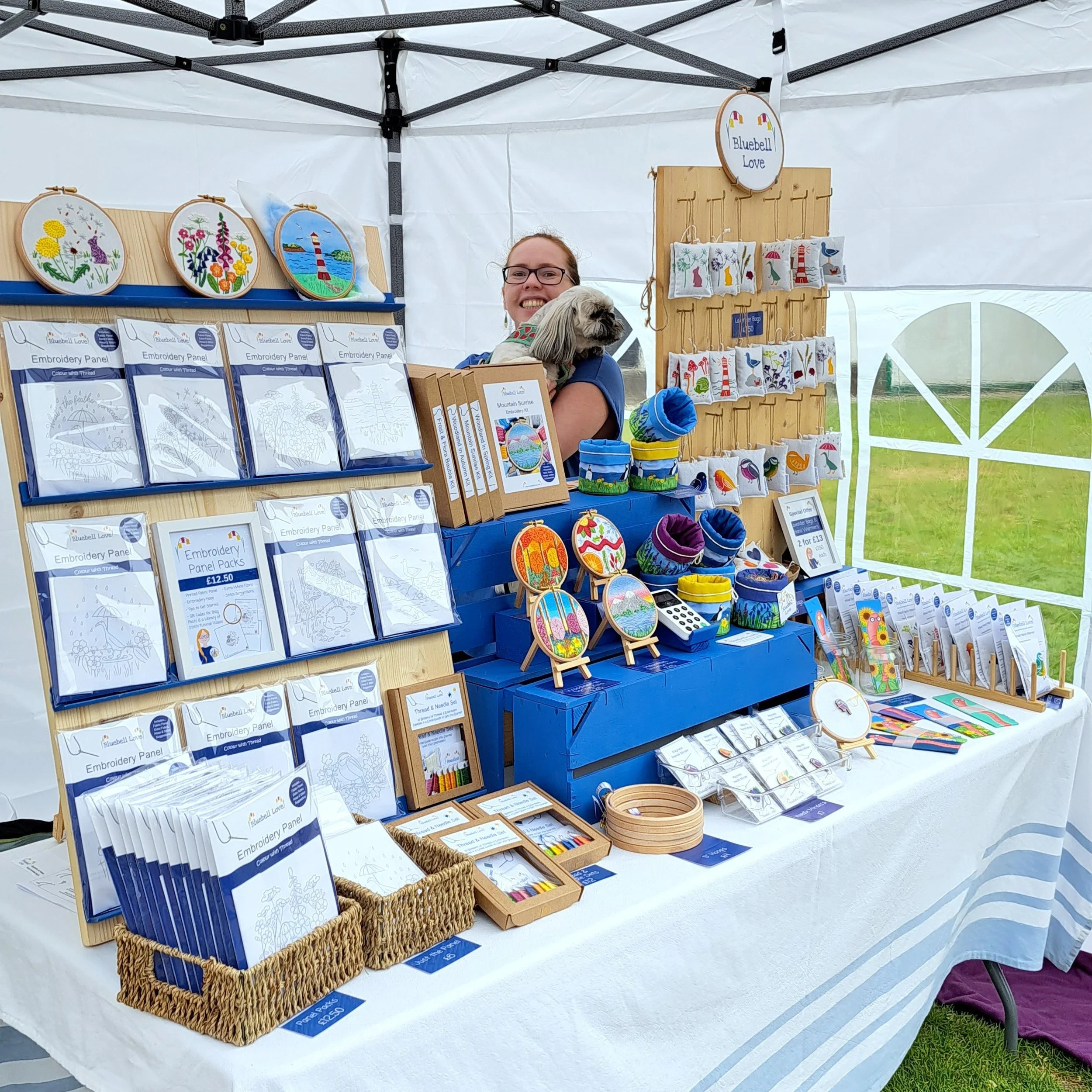 Liz from Bluebell Love at her craft stall, holding her dog Bessie behind a display of embroidery designs