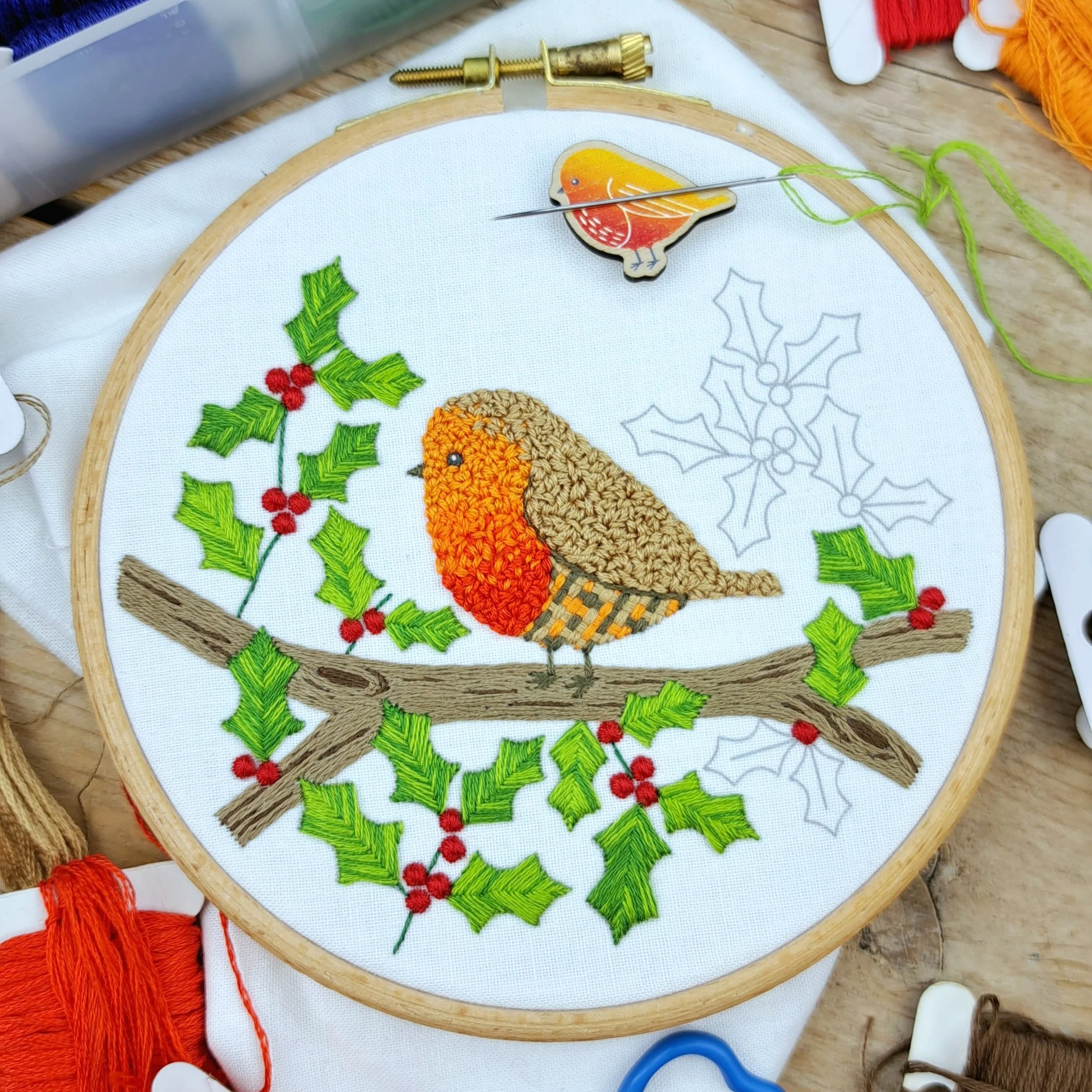 Partly completed embroidery of a robin on a holly branch, with just a few leaves, berries, and stems left to stitch.