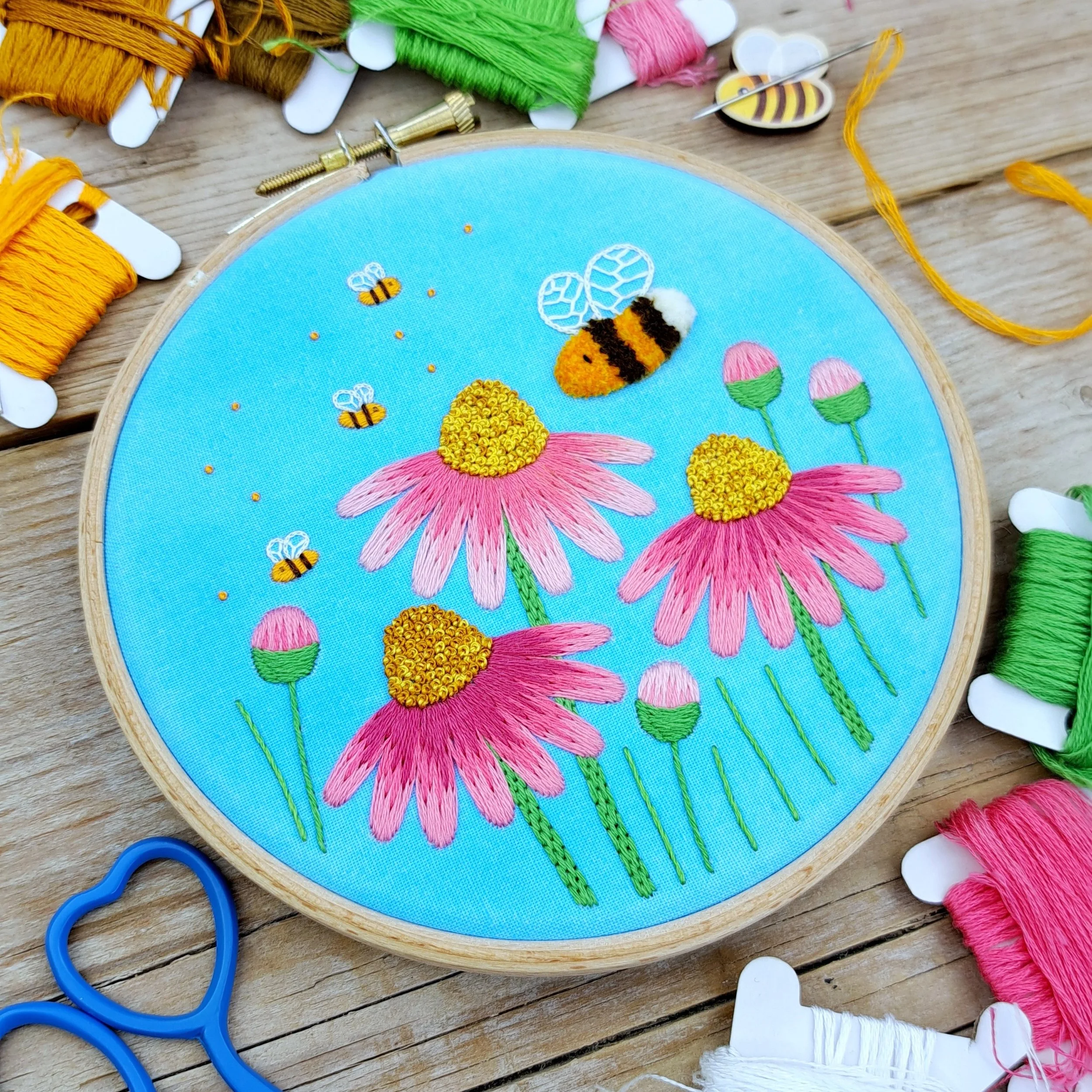 Finished embroidery of colourful pink flowers and buzzing bees on a blue watercolour painted background, displayed in an embroidery hoop.