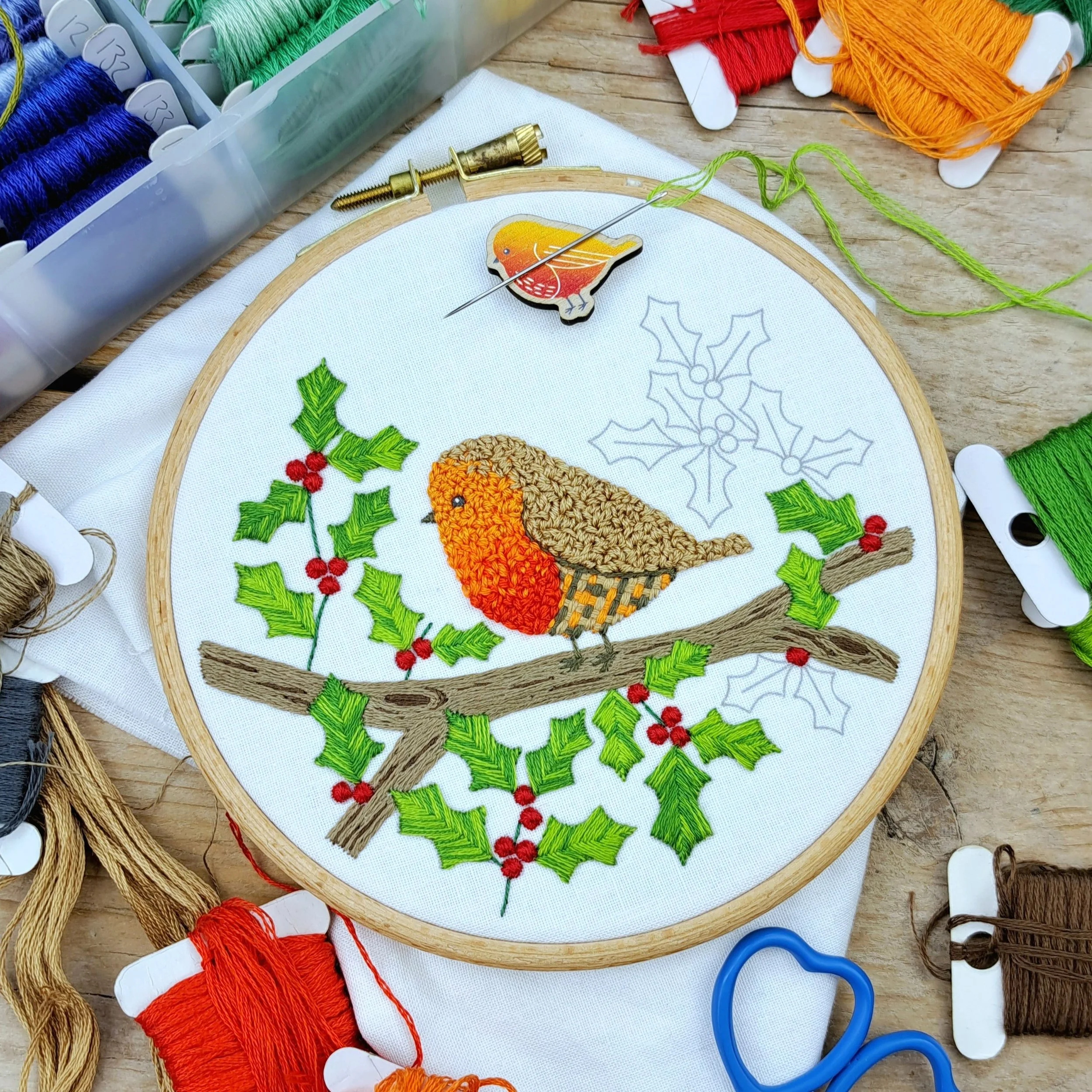 Mostly finished embroidery of a robin on a holly branch with berries, displayed in an embroidery hoop.