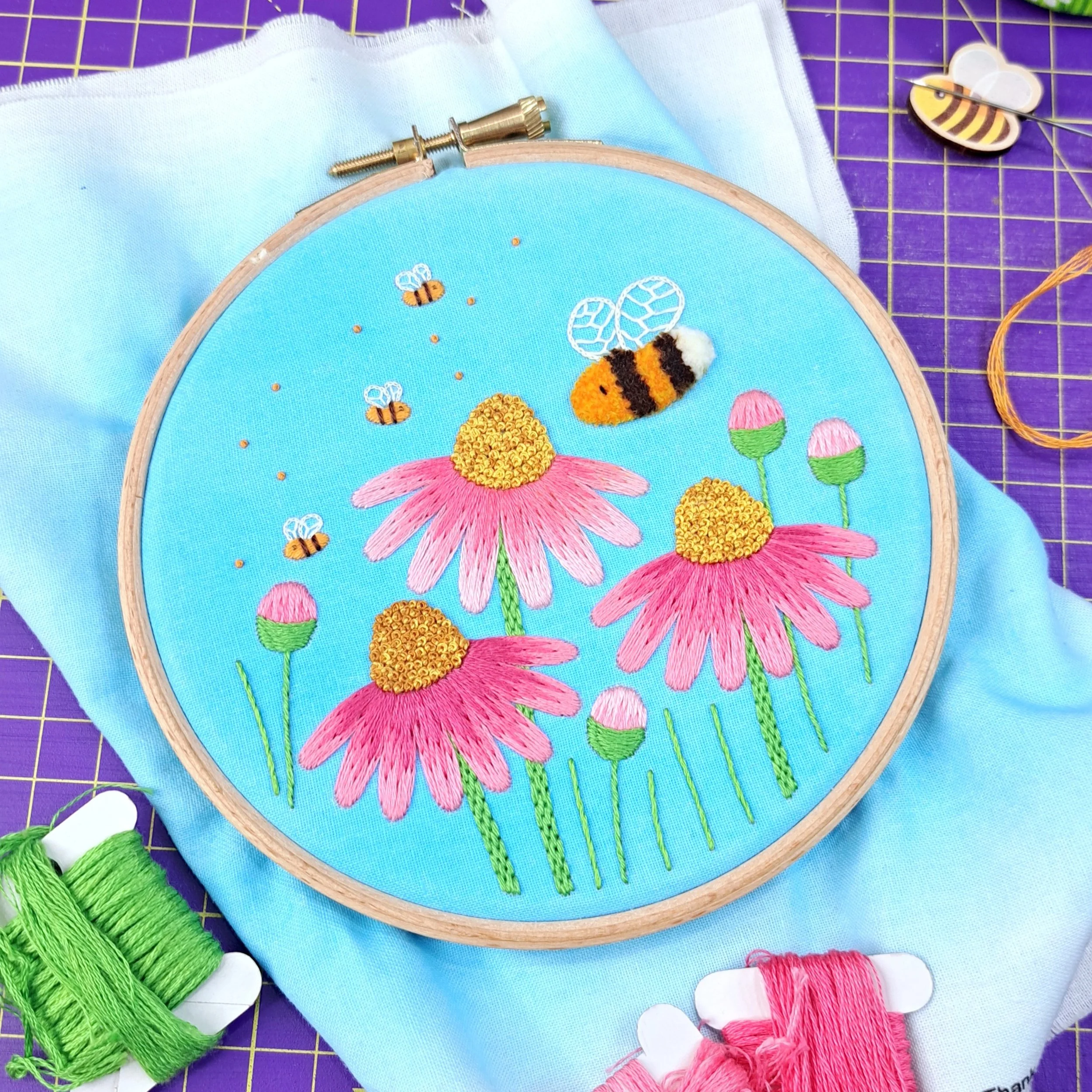 Finished embroidery of pink flowers, buds and flying bees with a blue watercolour painted background, displayed in an embroidery hoop.