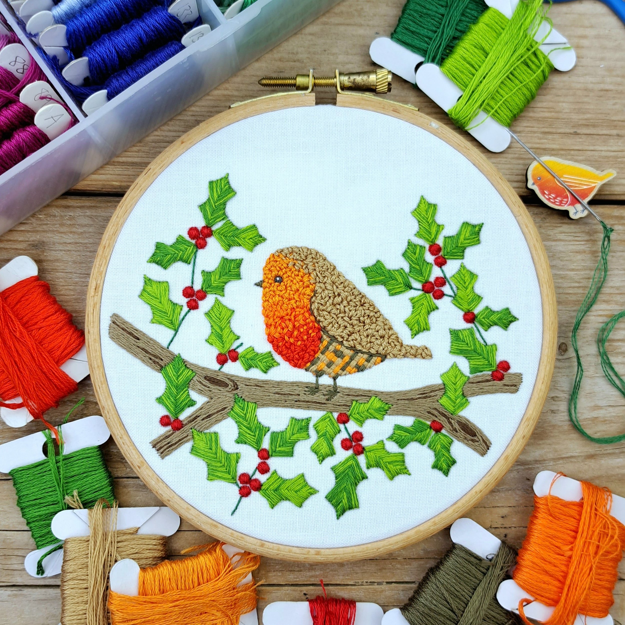 Finished embroidery of a robin perched on a holly branch, fully stitched with red berries and green holly leaves, displayed in an embroidery hoop.