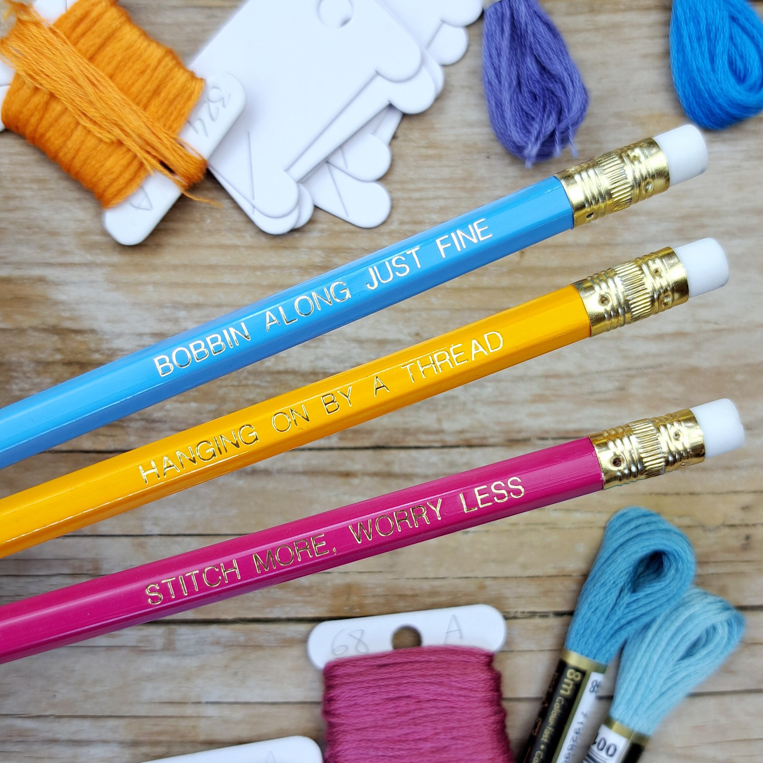 Set of three stitchy pencils showing the gold foil slogans and erasers, displayed above colourful embroidery threads