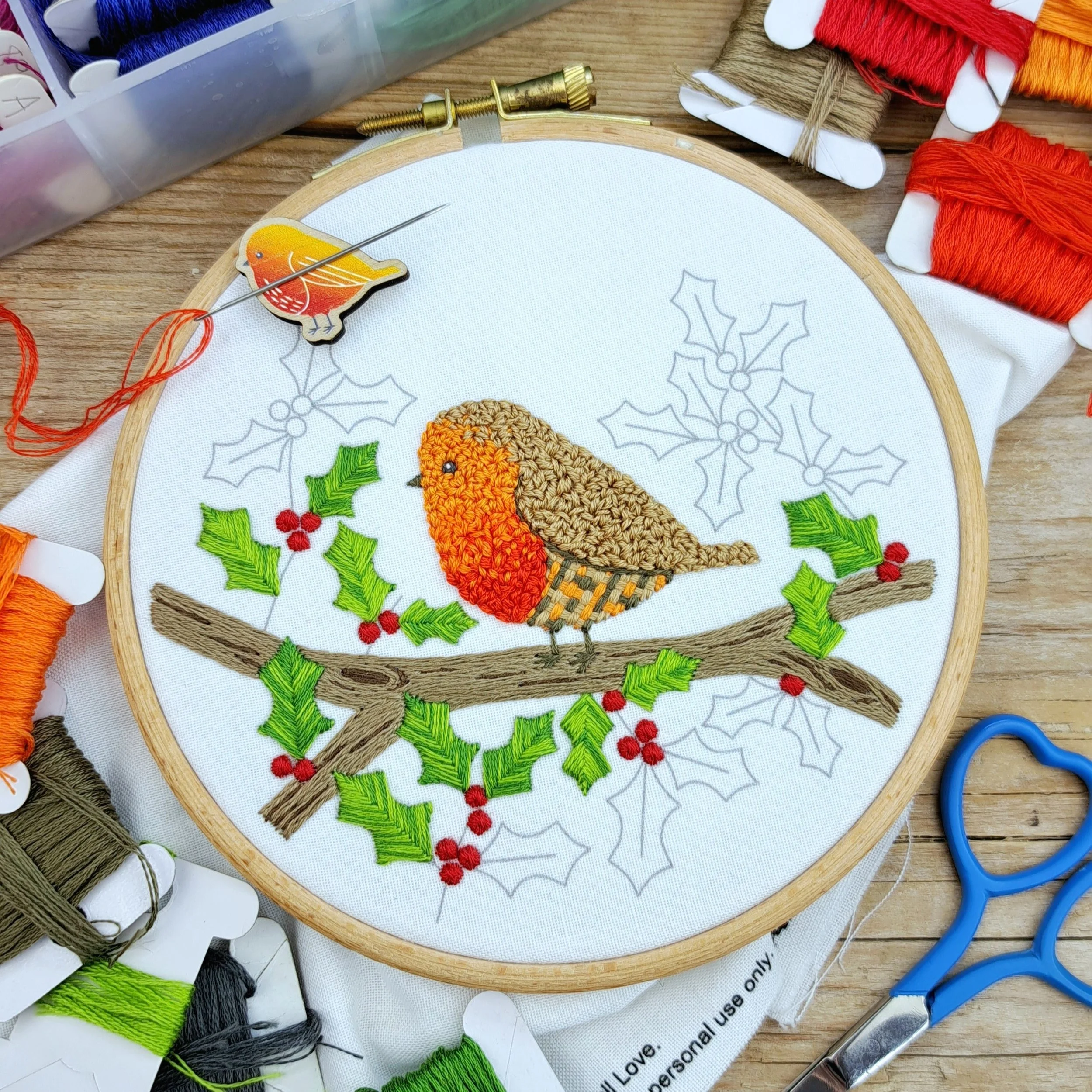 Partly completed robin on a holly branch, with the robin fully stitched and only a few leaves and berries remaining.
