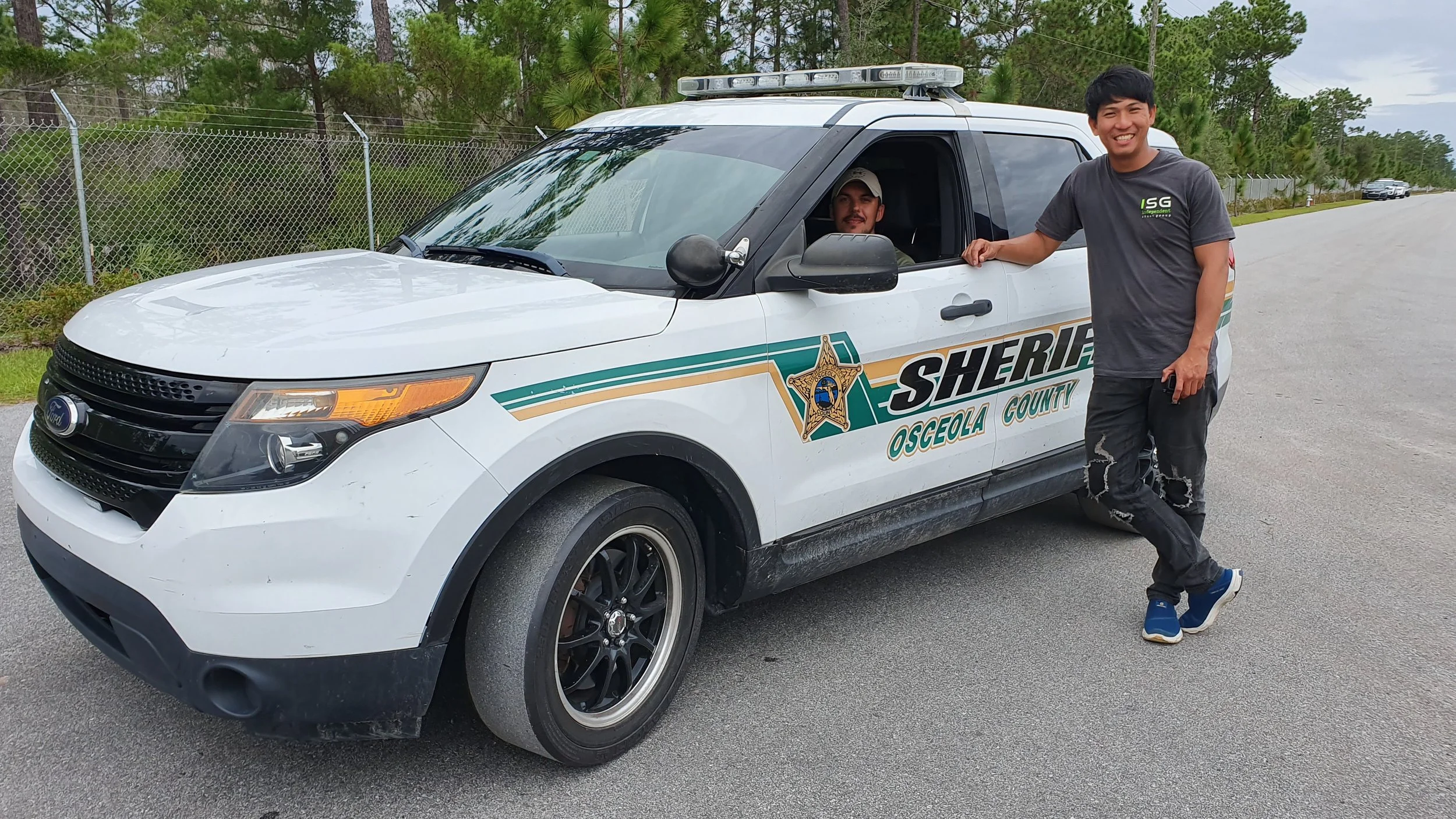 A young man (Kevin) smiling and standing with his arm on the door of a Osceola County sheriff SUV, with a deputy sitting inside the vehicle, on a paved road surrounded by trees.