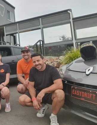 Two men and a woman posing outdoors near a gray sports car with an EasyDrift sign at a racetrack circuit. Charles.