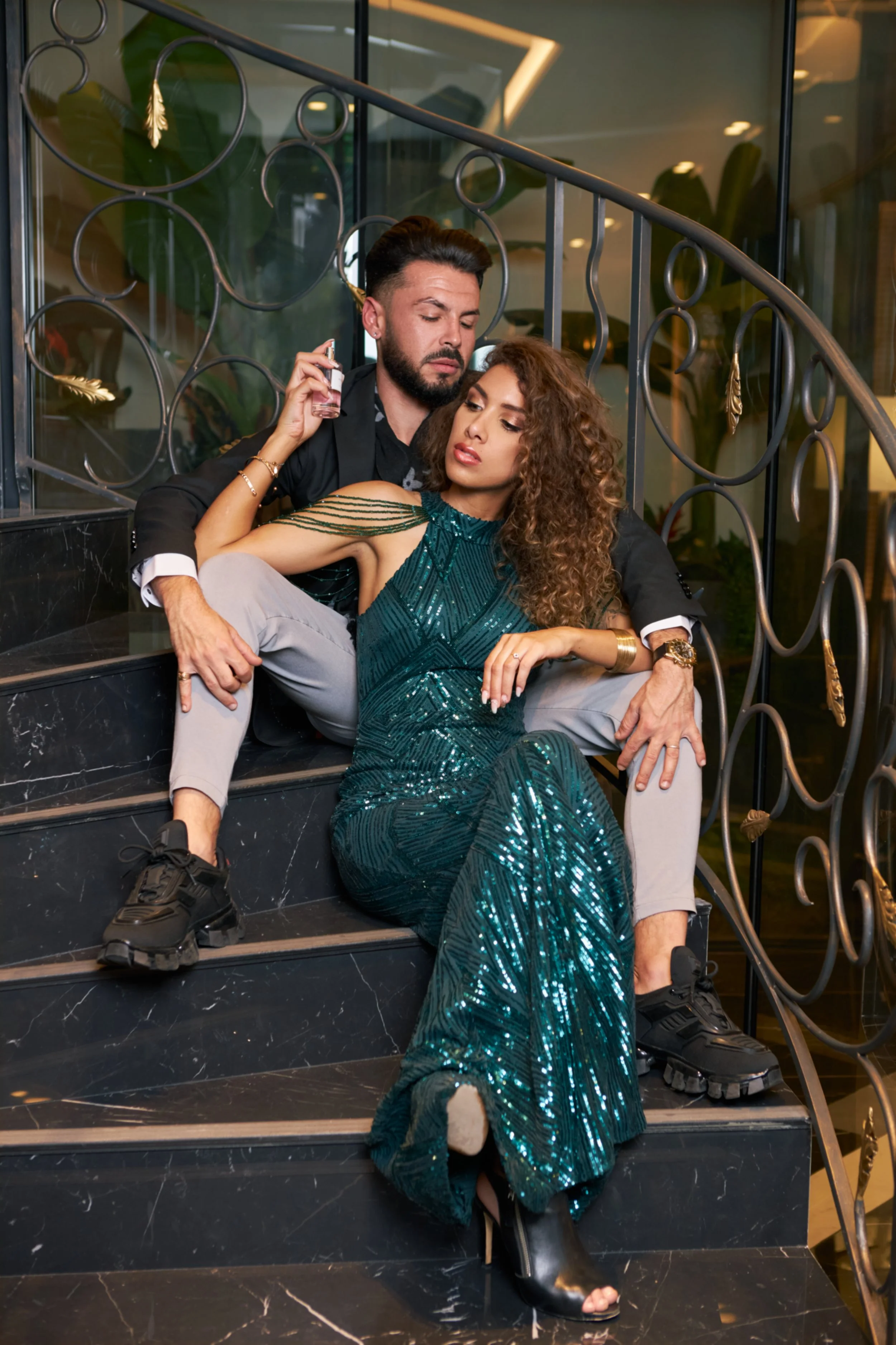 Un couple pose assis dans un escalier intérieur décoré, la femme portant une robe verte à sequins et l’homme un costume noir.